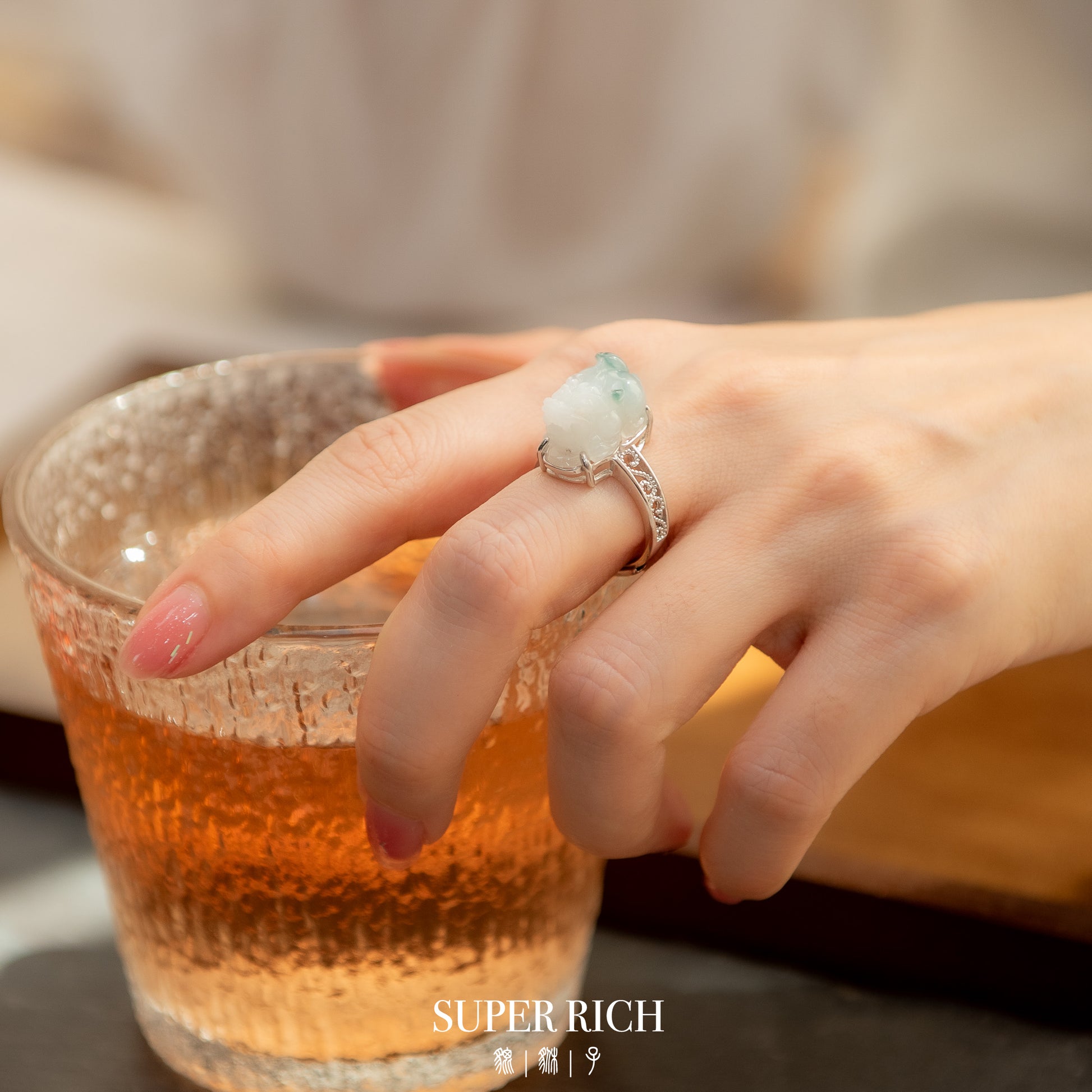 Hand wearing a ring with a large gemstone holding a glass of amber liquid, blurred background