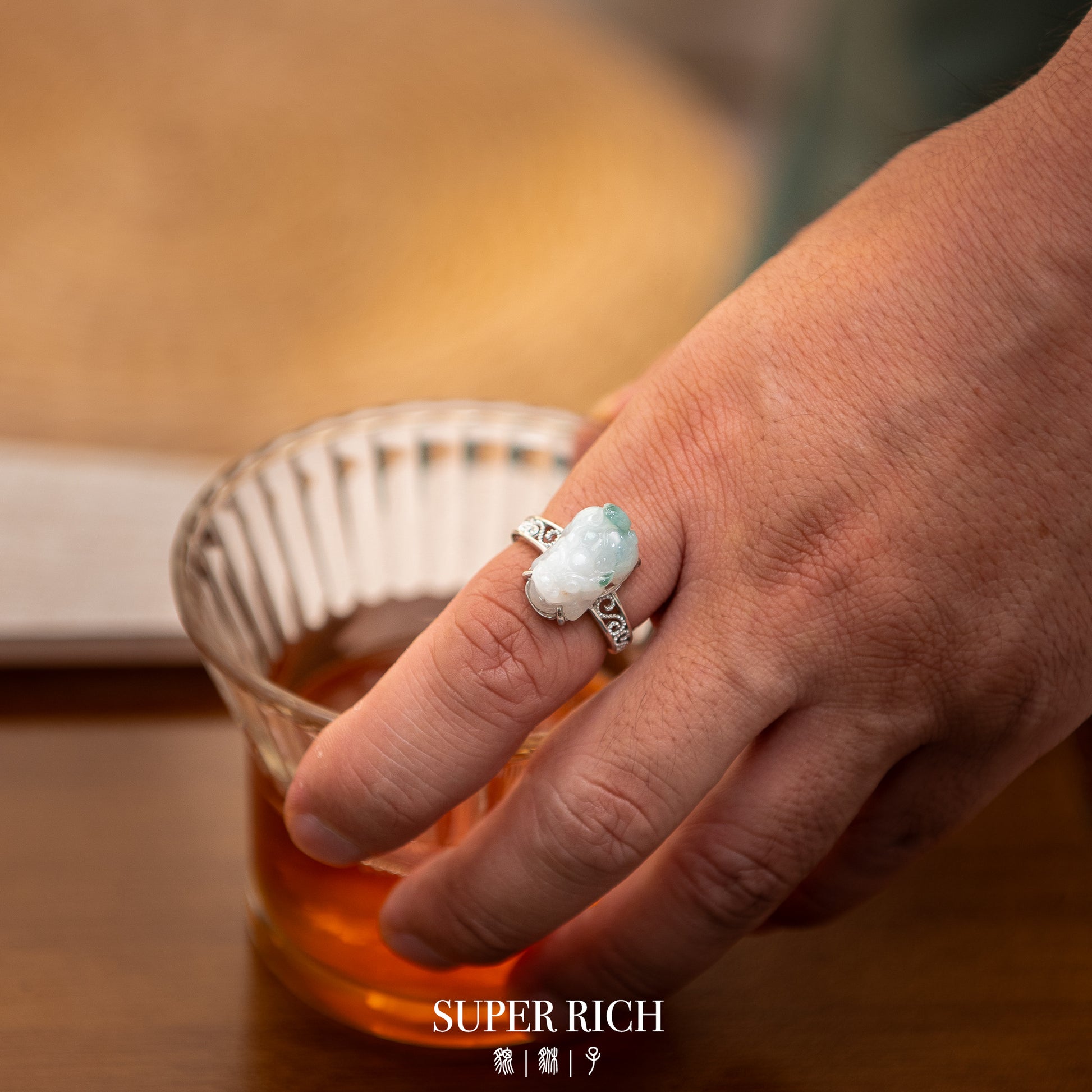 Hand holding a glass of amber liquid with a decorative ring, blurred background