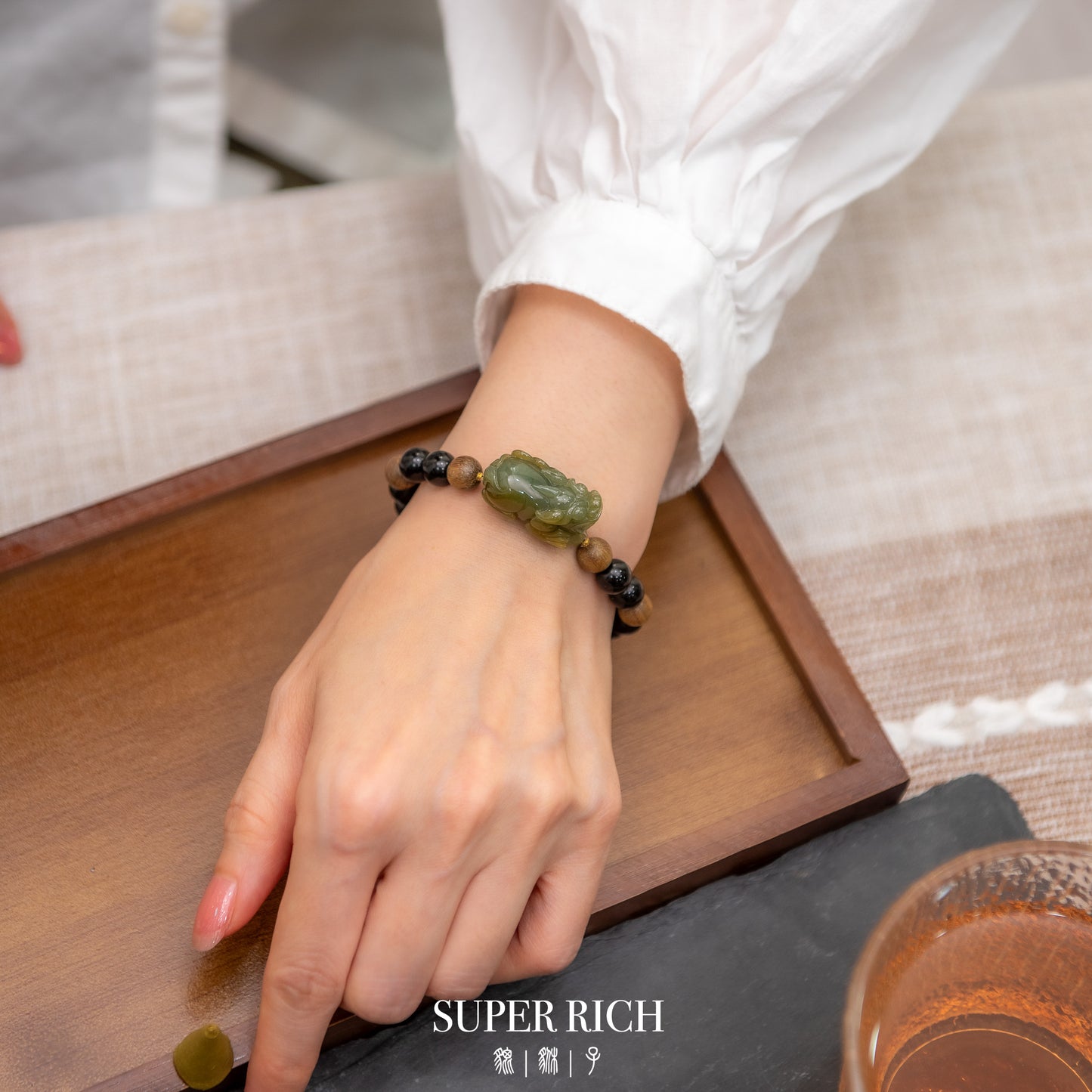 Hand wearing a bracelet with green beads on a wooden surface, with 'SUPER RICH' branding.