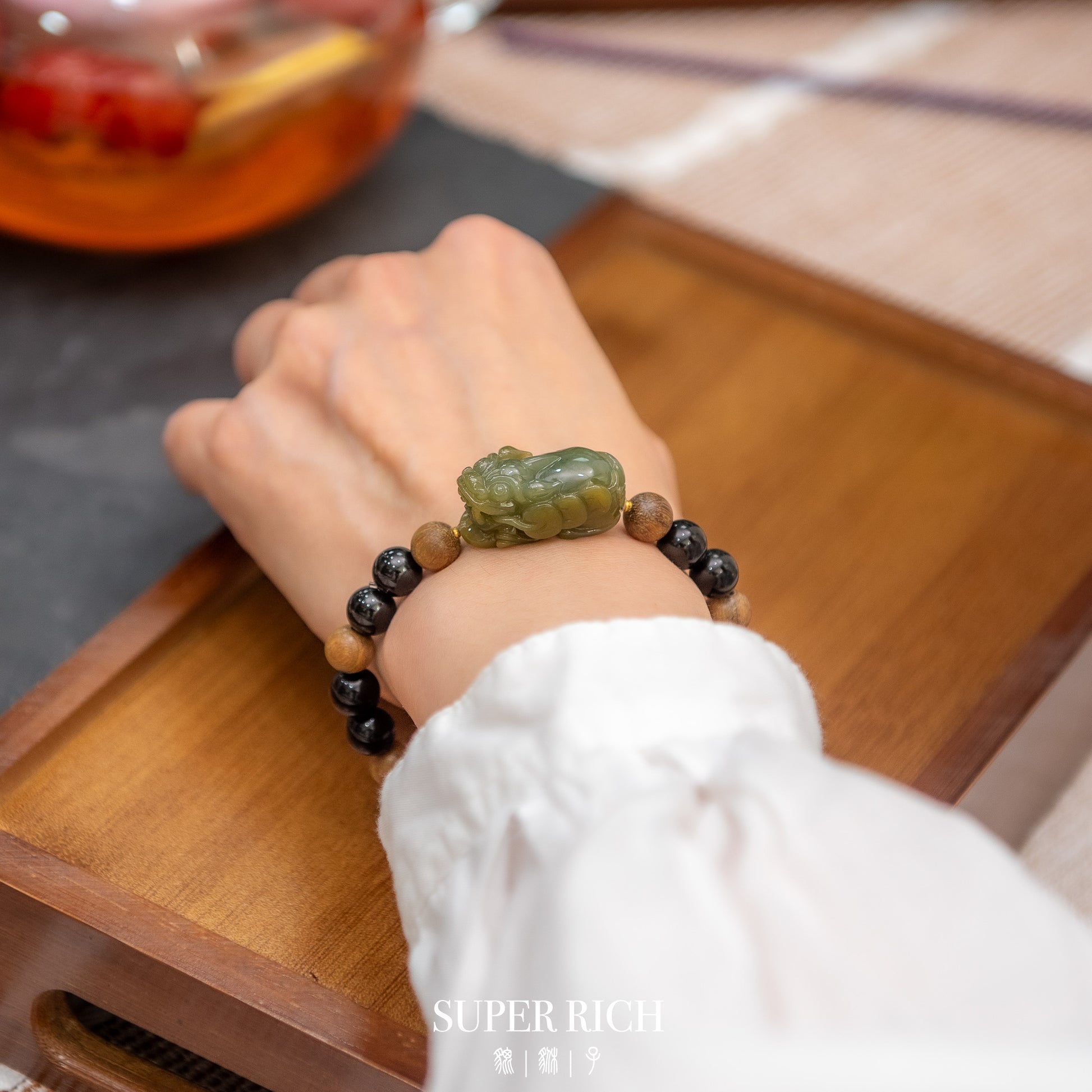 Hand wearing a bracelet with a green stone on a wooden surface, with 'Super Rich' branding.