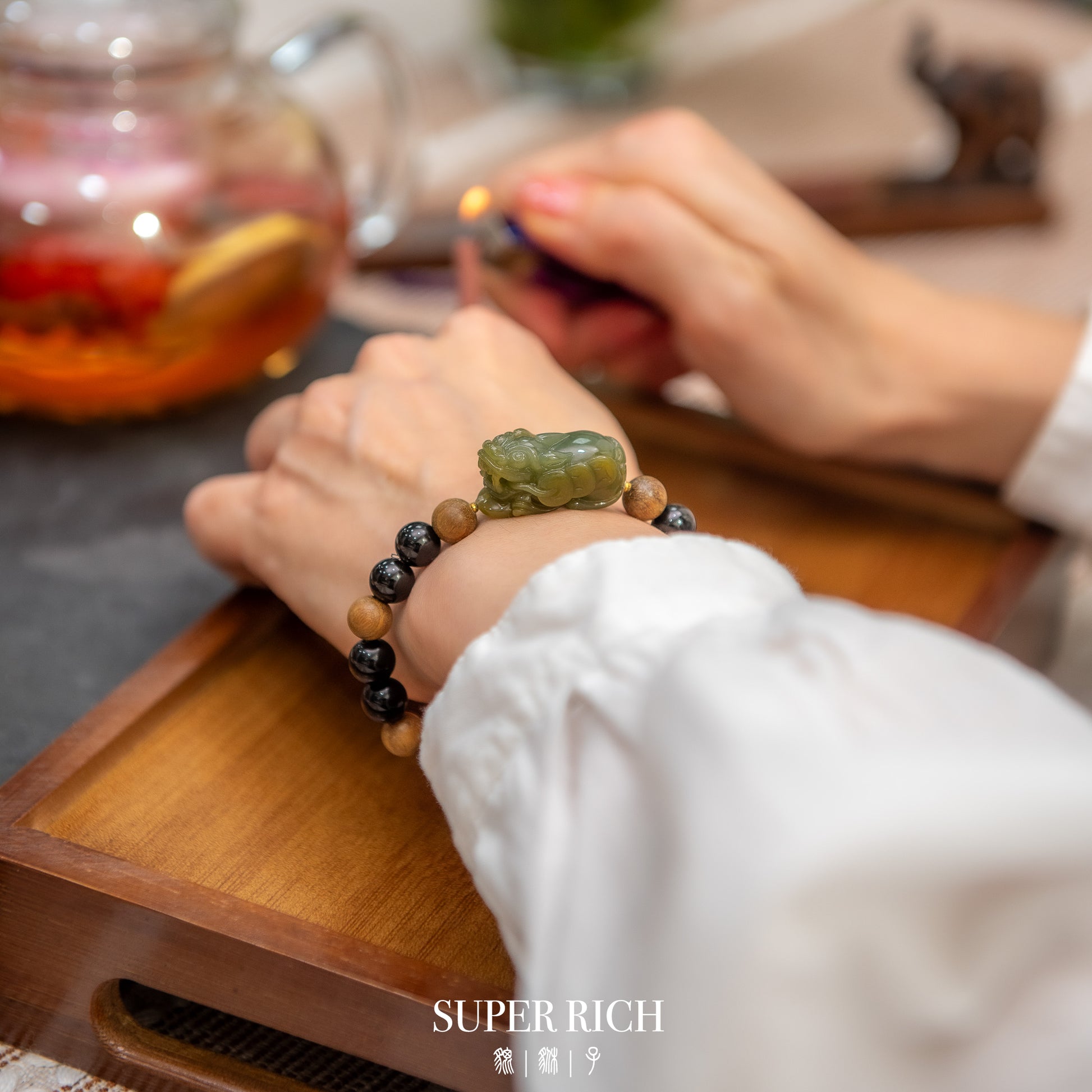 Person wearing a bracelet with a green stone on a wooden tray, with 'SUPER RICH' branding.