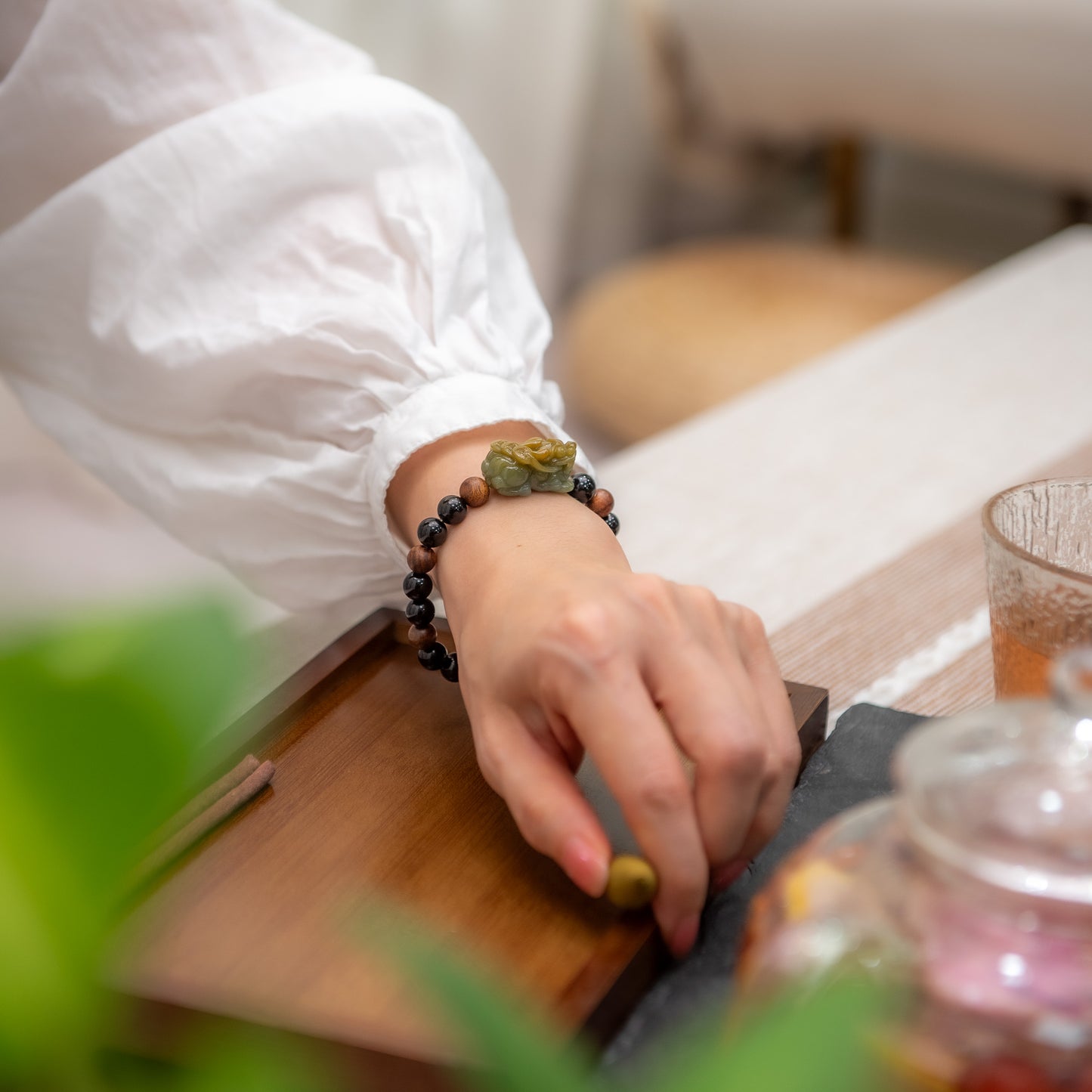 Person wearing a bracelet with a small figurine, sitting at a table with a teapot and cup.