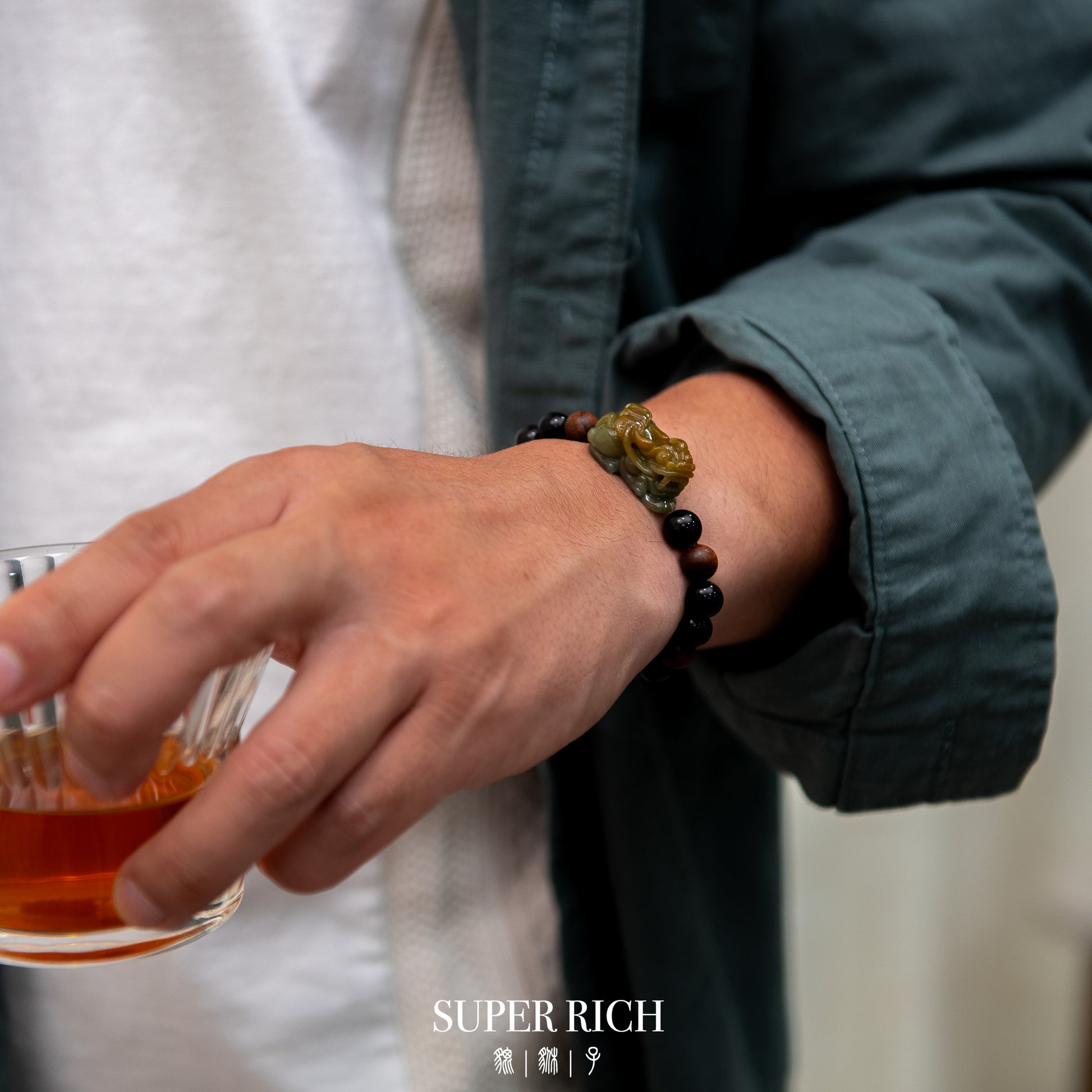Hand holding a glass of amber liquid with a bracelet, wearing a green jacket, on a neutral background.