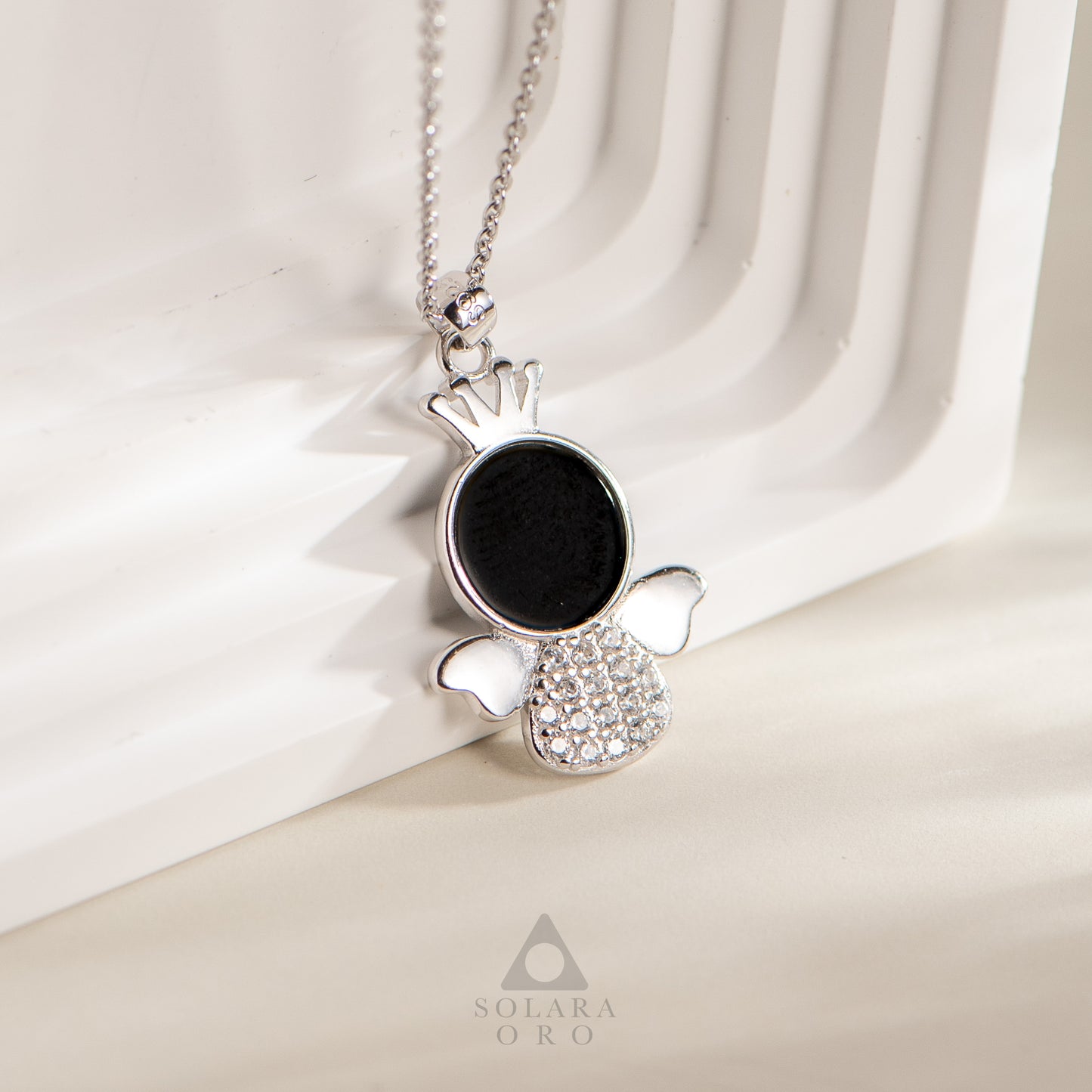 Silver pendant with black stone and clear stones on a white fabric background, featuring the brand 'Solaara Oro'.