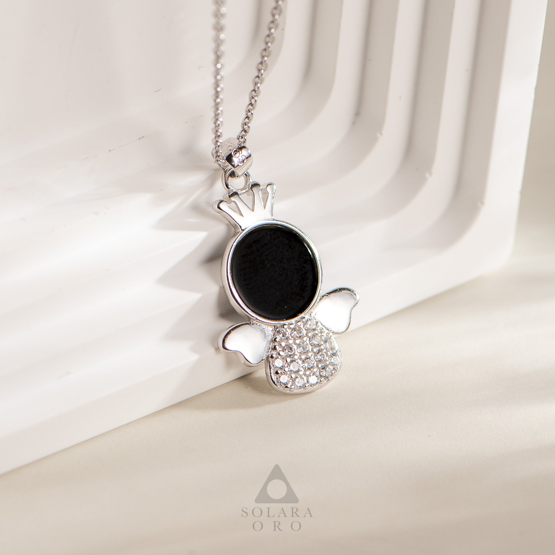 Silver pendant with black stone and clear stones on a white fabric background, featuring the brand 'Solaara Oro'.