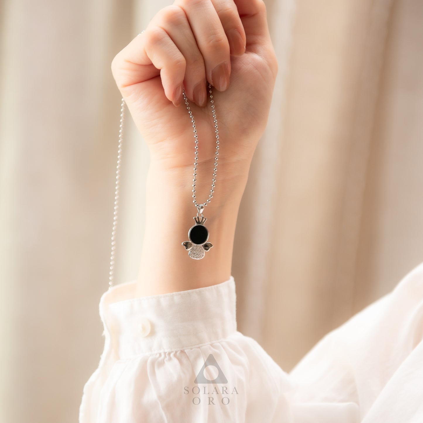 Hand wearing a silver necklace with a black pendant against a beige background