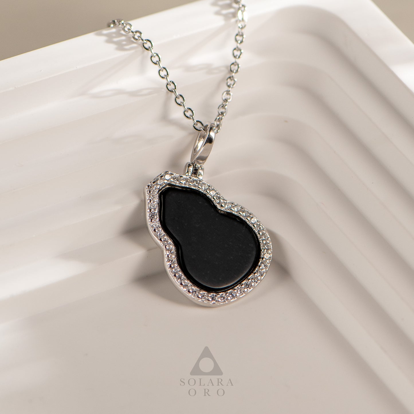 Necklace with a black pendant and silver chain on a beige fabric background, featuring the brand 'Soleara Oro'.