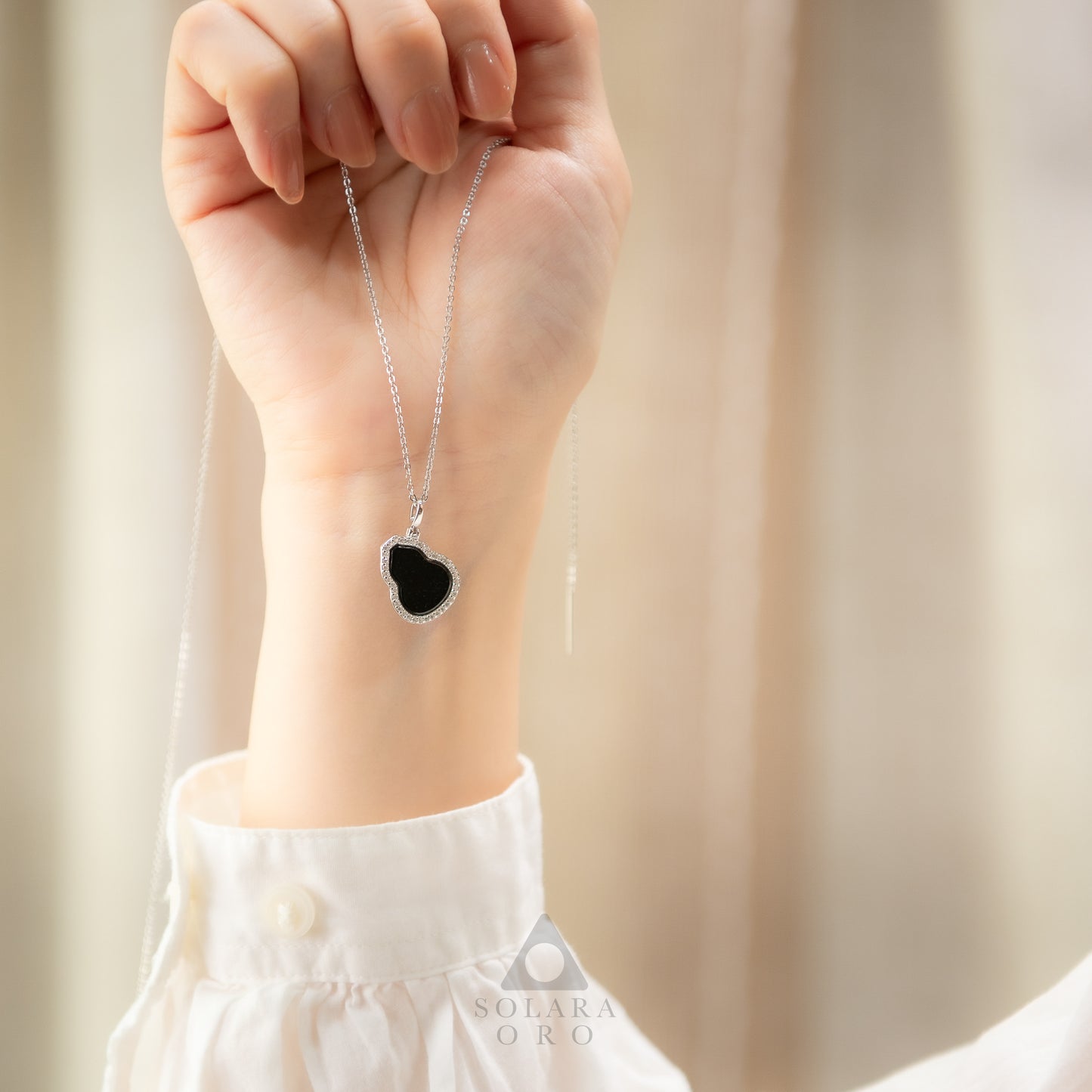 Hand wearing a necklace with a black heart pendant against a neutral background