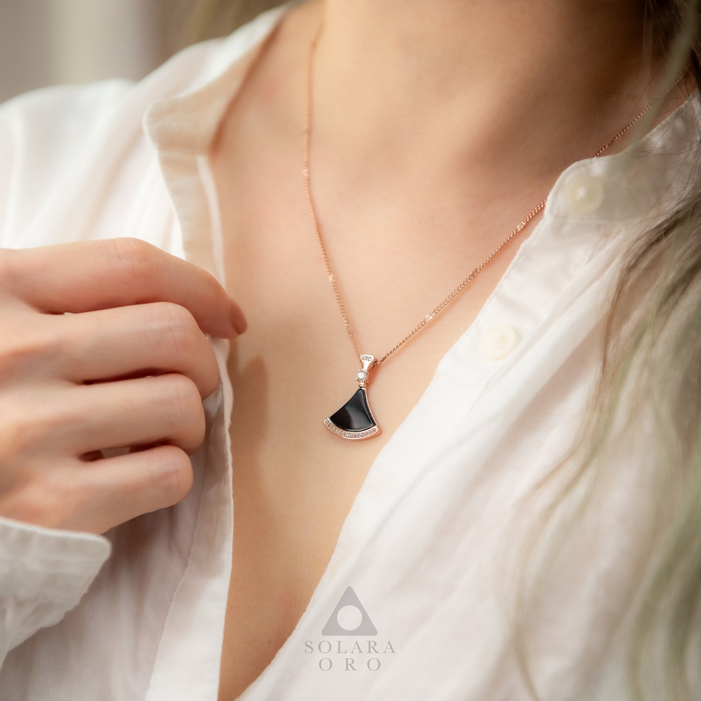 Person wearing a necklace with a black pendant on a blurred background, featuring the brand 'Solaara Oro'.