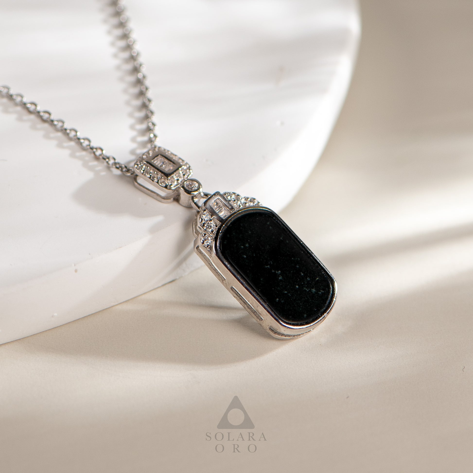 Necklace with a black pendant and silver chain on a white fabric background, featuring the Solara Oro brand.