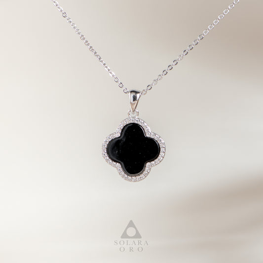 Black clover-shaped pendant on a silver chain with 'Solara Oro' branding against a neutral background.