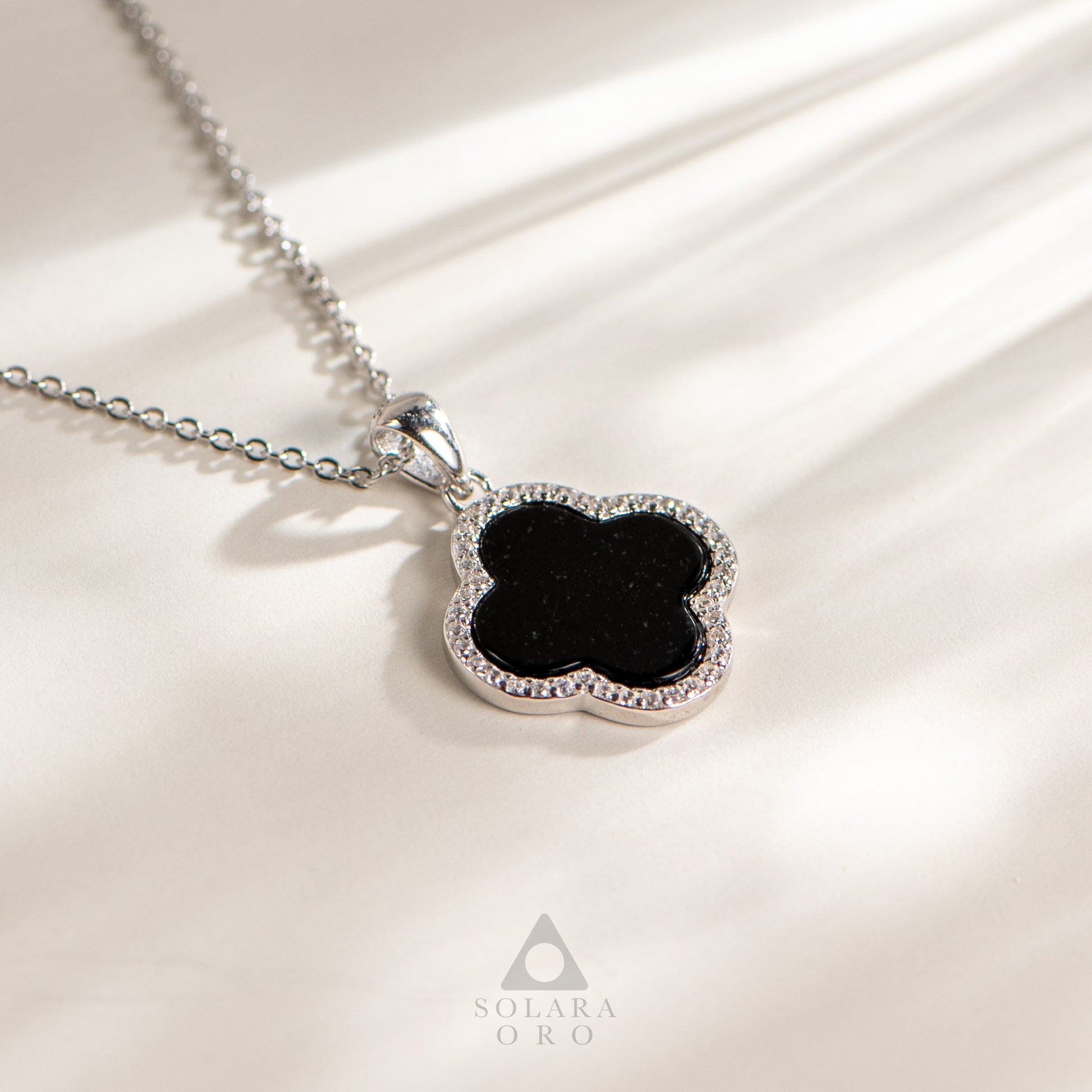 Silver necklace with a black clover pendant on a beige fabric background, featuring the Solara Oro brand.