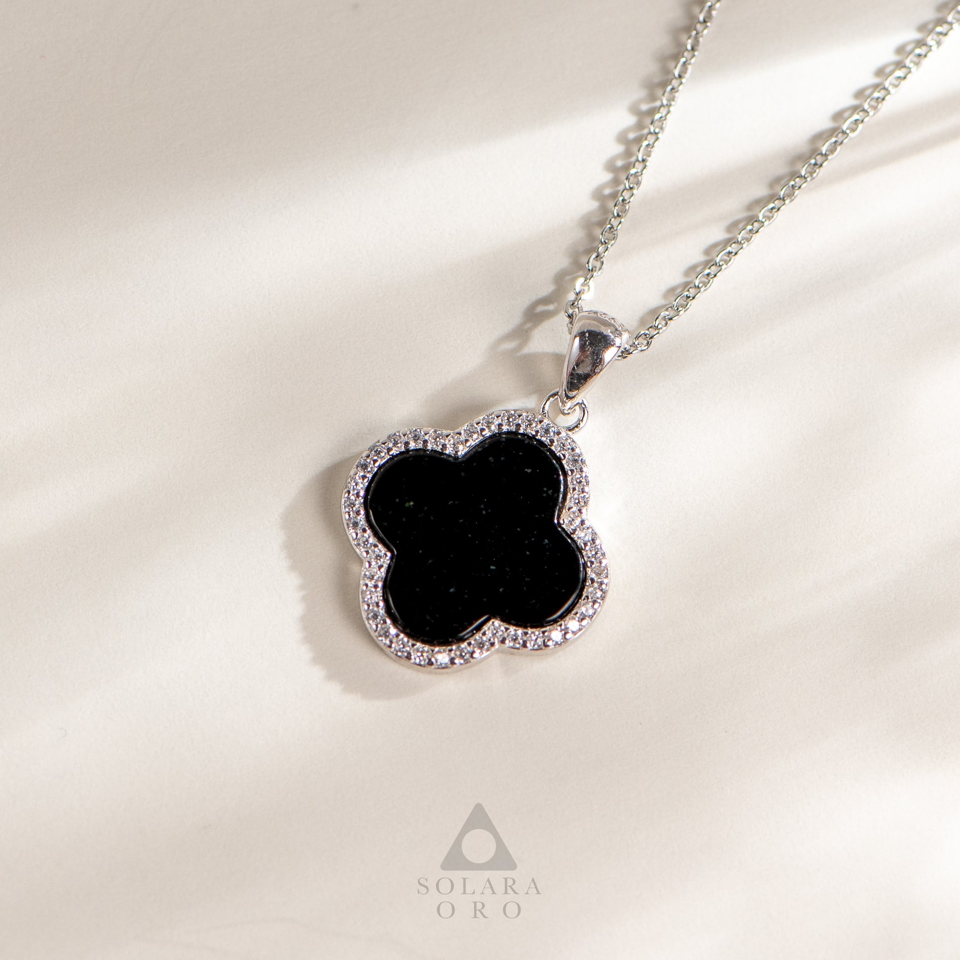 Black clover-shaped pendant on a chain with 'Soleara Oro' branding on a light background
