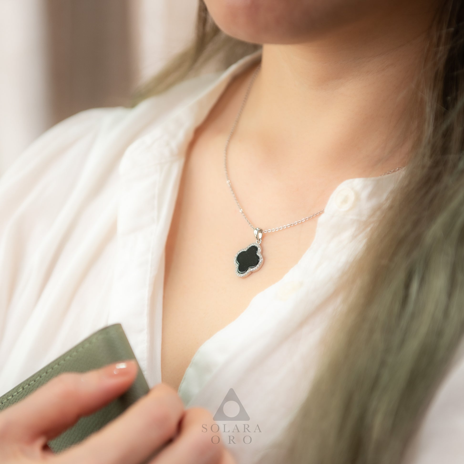 Person wearing a necklace with a clover pendant, holding a green card, against a neutral background.