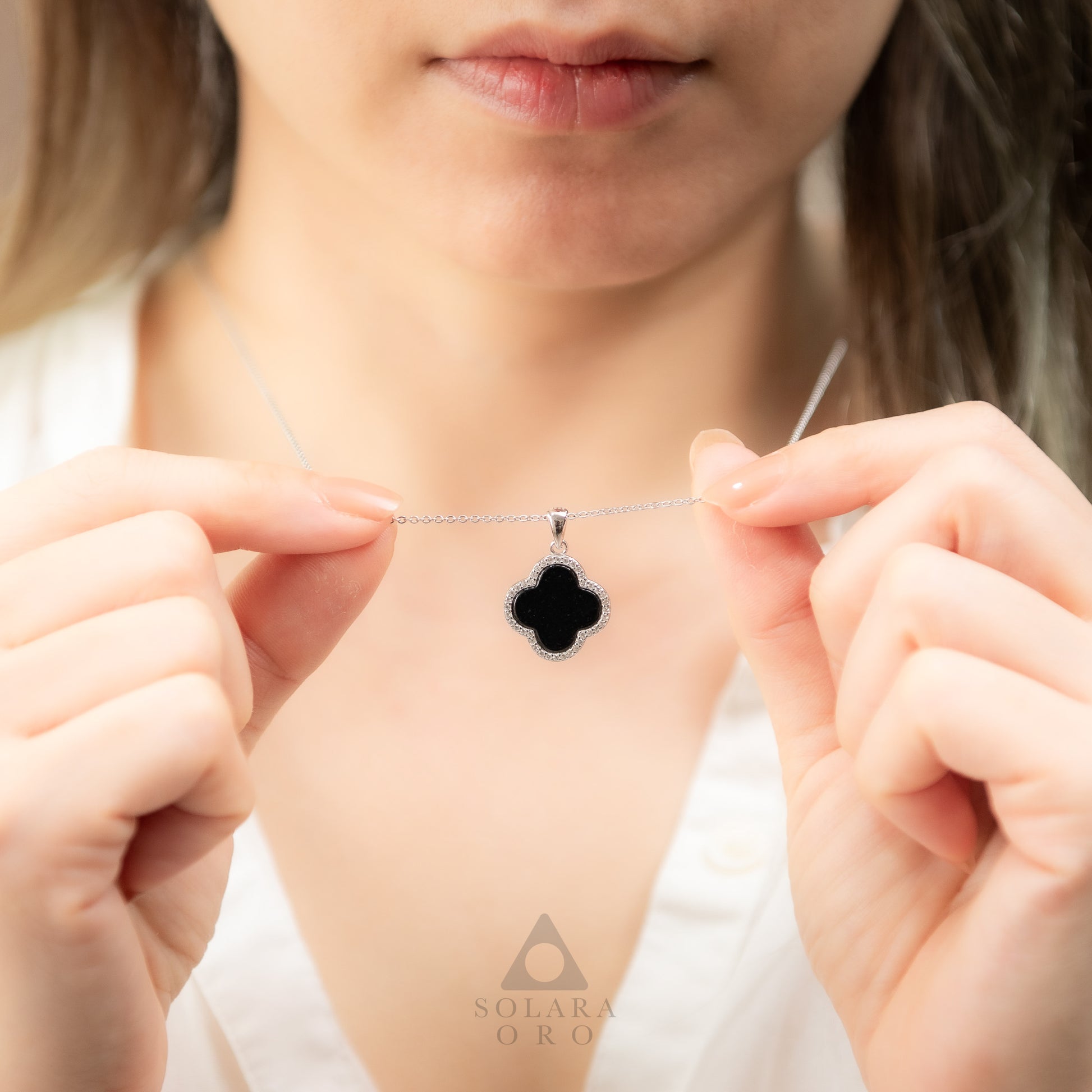 Person holding a necklace with a clover-shaped pendant, brand 'Solaire Oro' visible.
