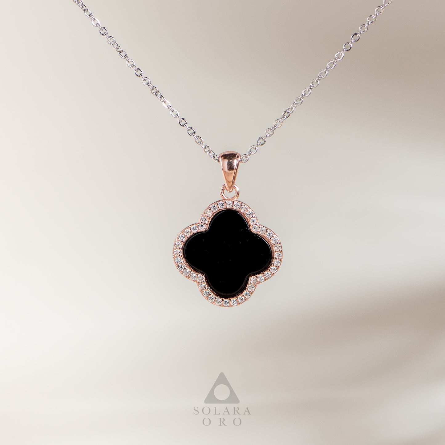 Clover-shaped pendant with black center and rose gold frame on a chain against a light background.