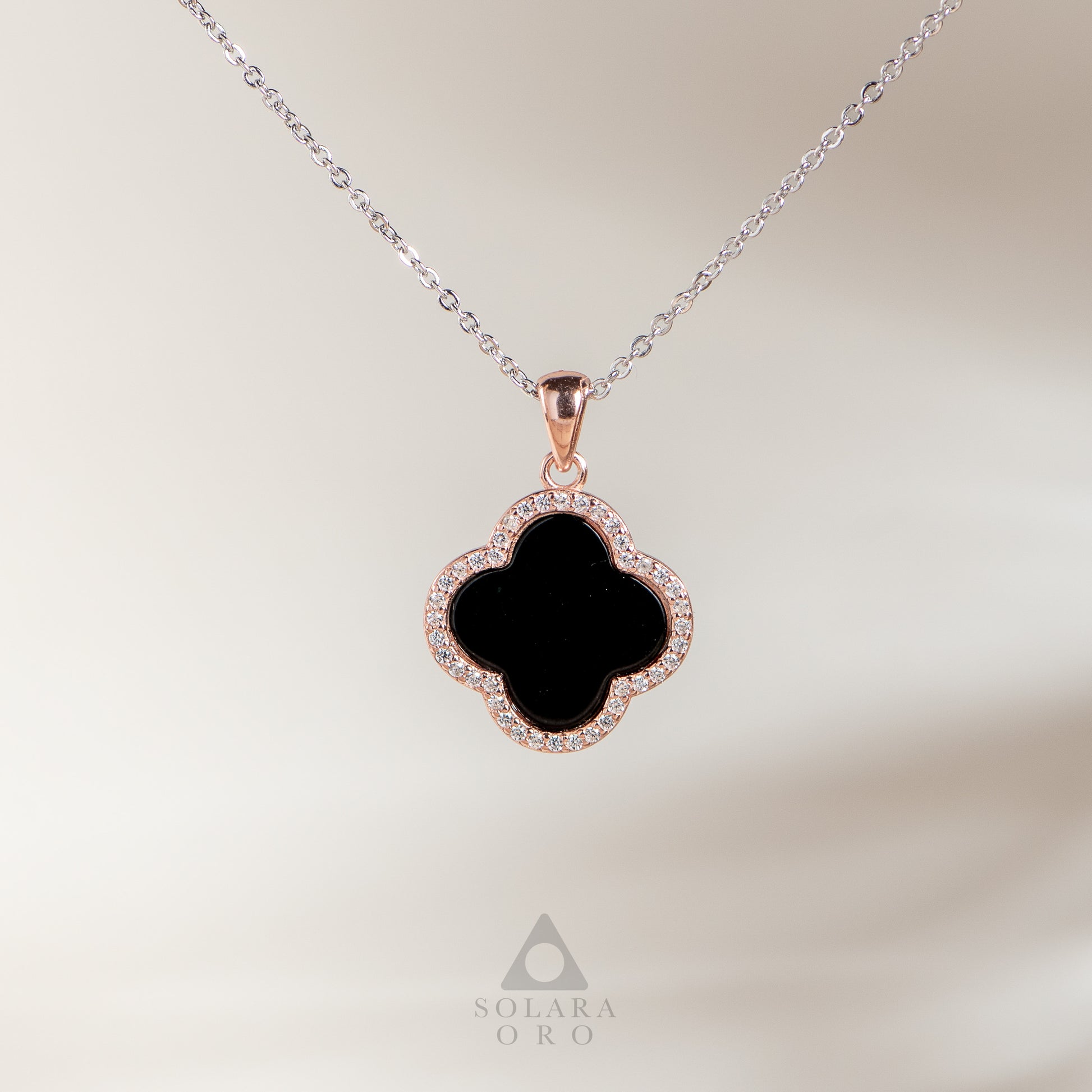 Clover-shaped pendant with black center and rose gold frame on a chain against a light background.