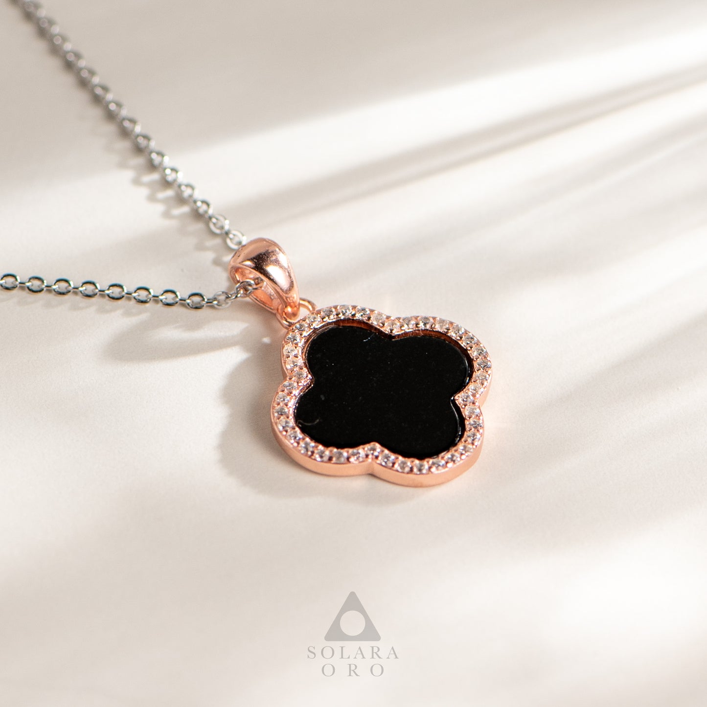 Necklace with a black clover pendant and rose gold border on a white fabric background, featuring the Solara Oro brand.
