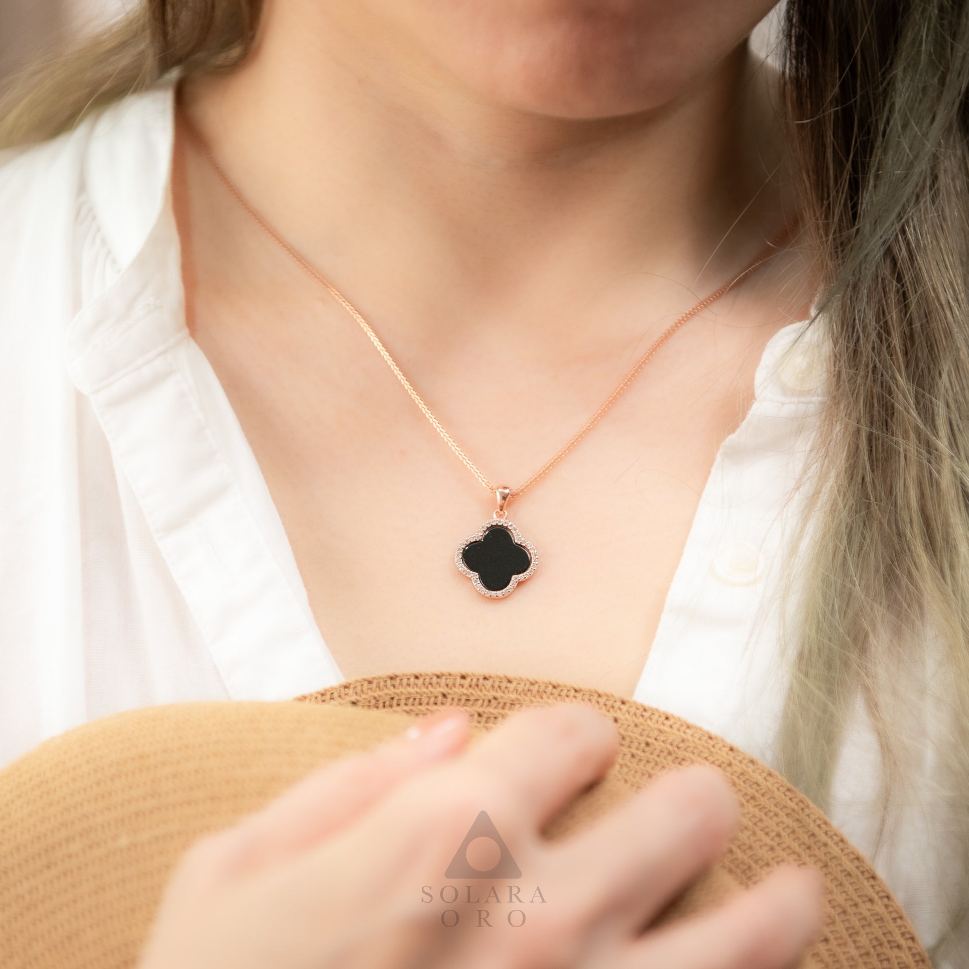Person wearing a necklace with a black clover pendant, brand 'Sulara Oro' visible.