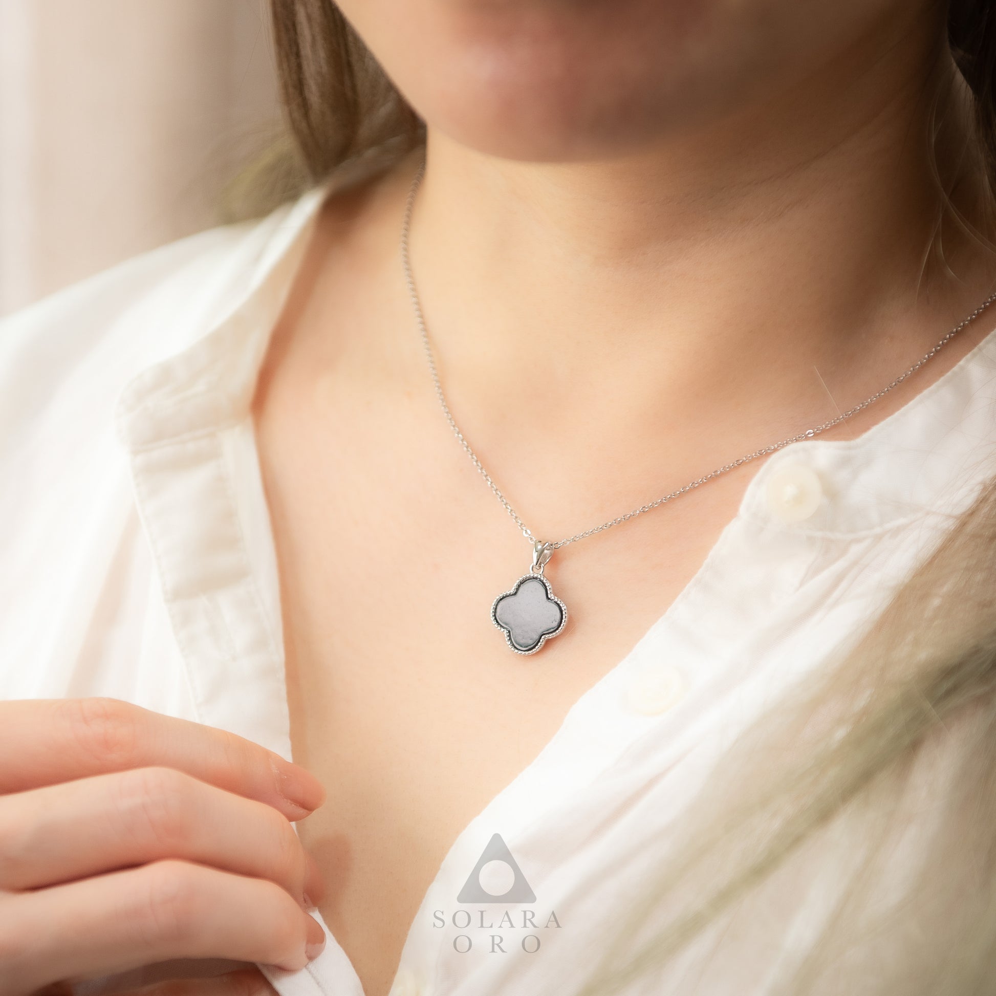 Person wearing a silver necklace with a clover-shaped pendant, brand 'Solara Oro' visible.