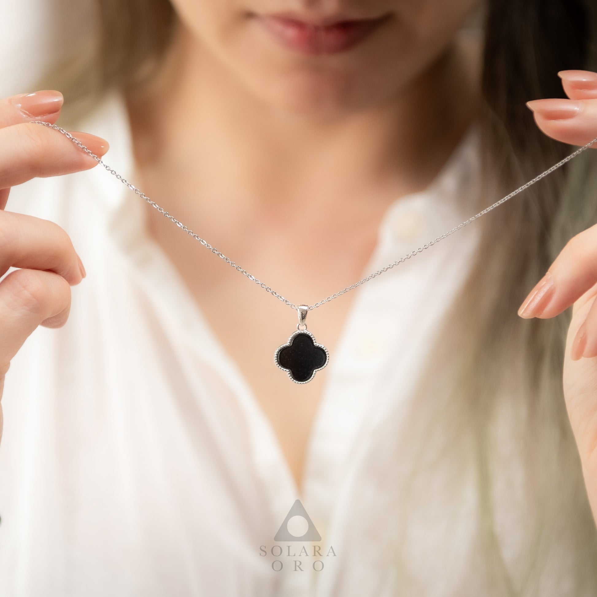 Black clover-shaped pendant on a silver chain held by a person wearing a white top.