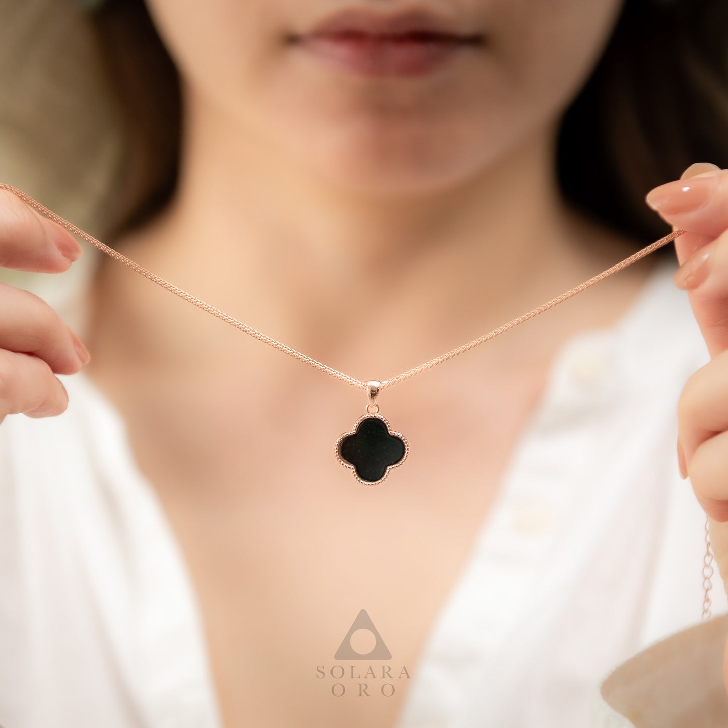 Person holding a necklace with a clover-shaped pendant, brand 'SOLARA ORO' visible.