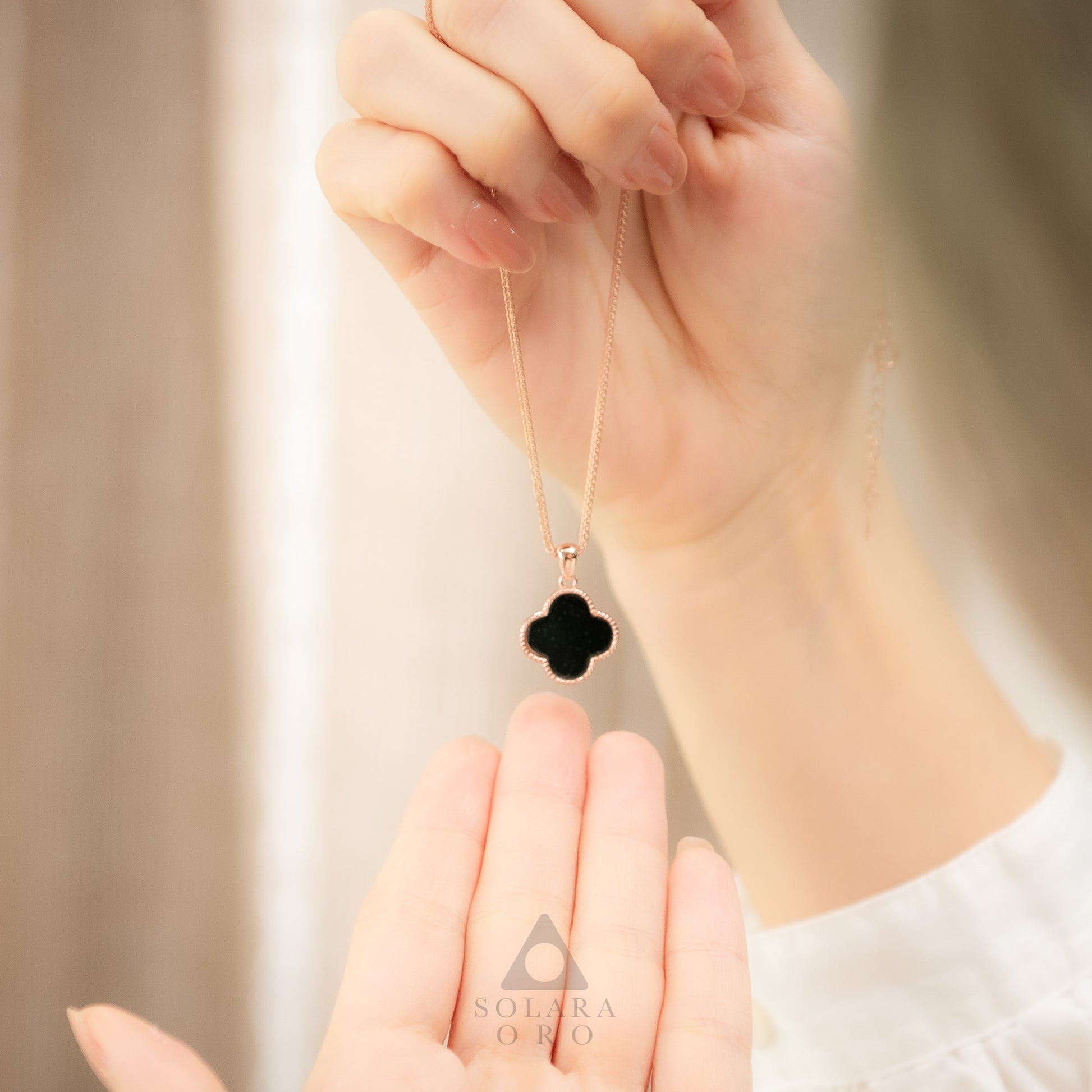Hand holding a necklace with a black clover pendant against a neutral background