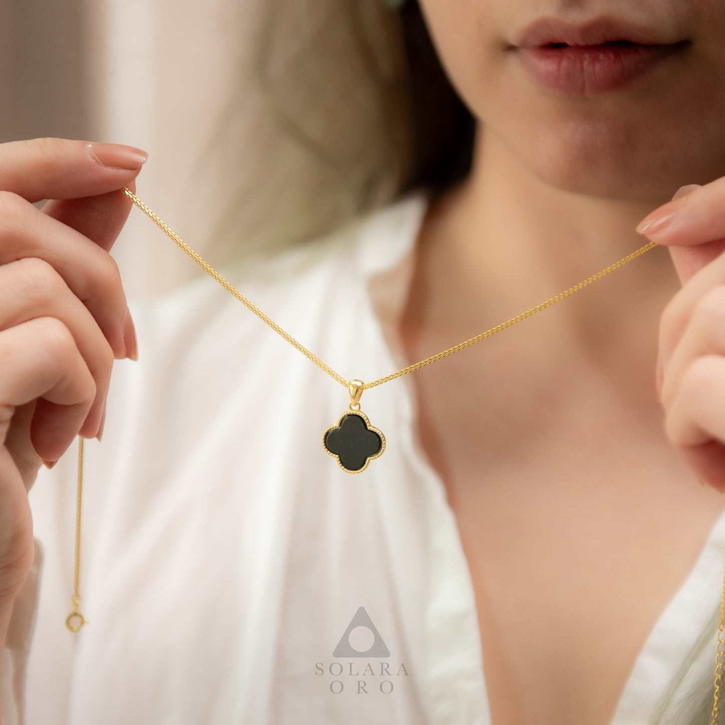 Gold necklace with a clover-shaped pendant held by a person, brand 'Solerara Oro' visible.