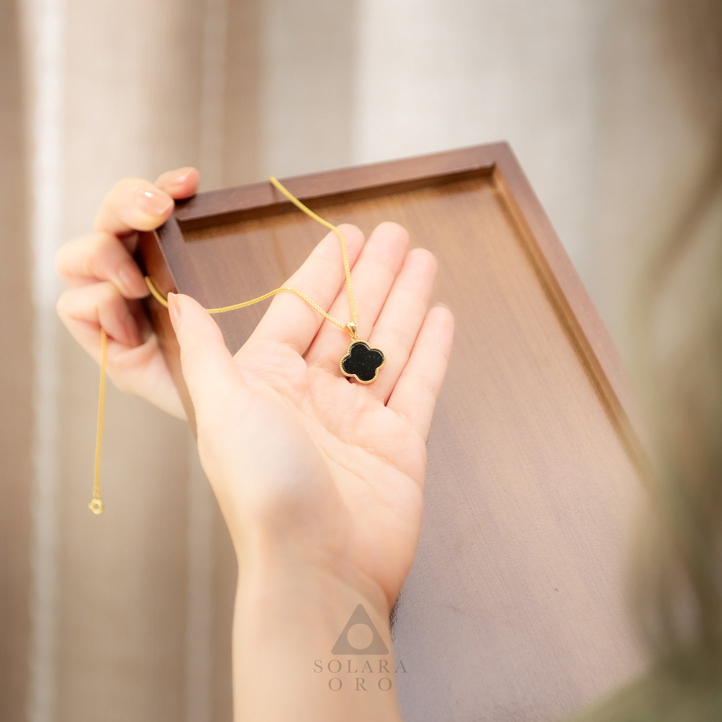 Hand holding a gold necklace with a black clover pendant in front of a wooden frame.