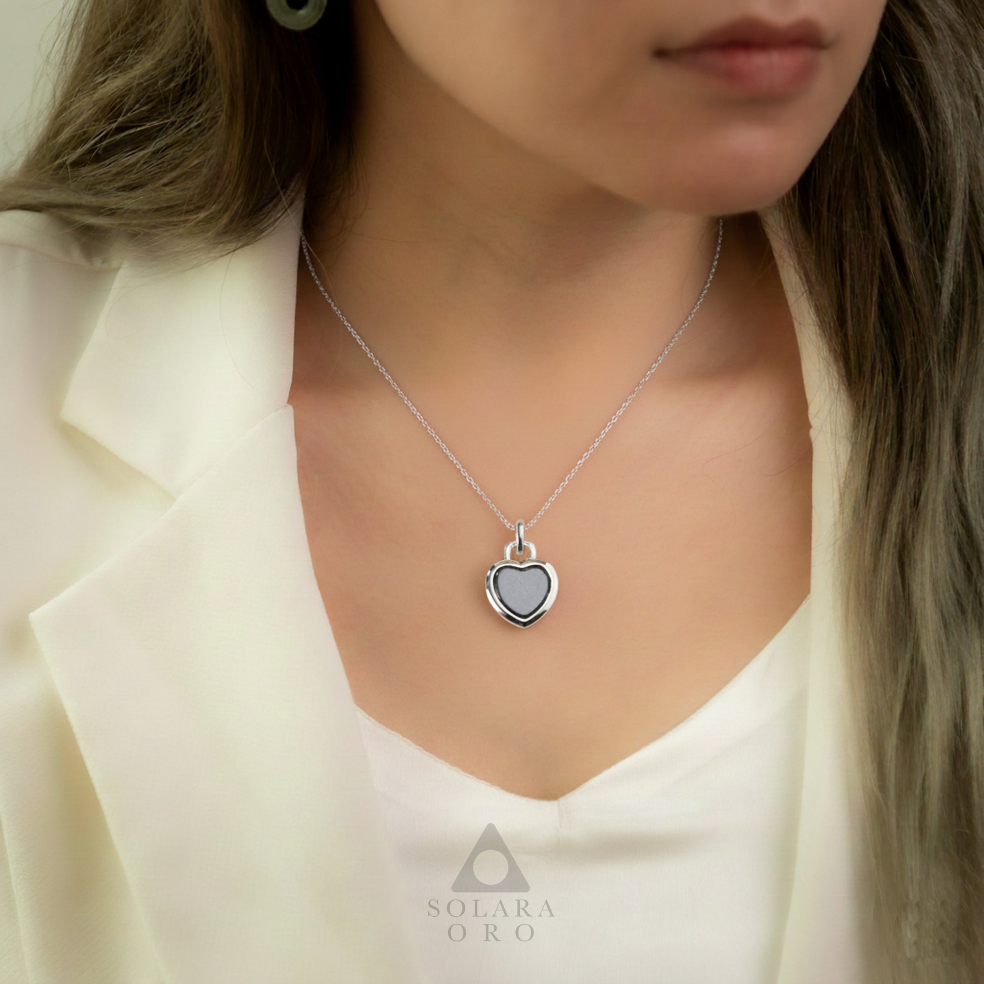 Woman wearing a silver necklace with a heart-shaped pendant, branded 'Solara Oro'.