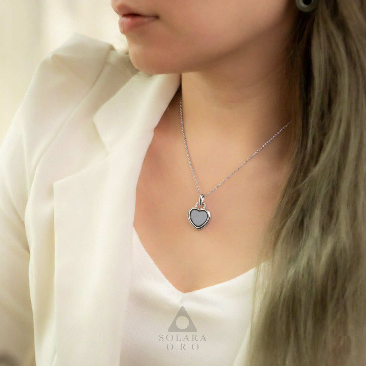 Woman wearing a silver necklace with a heart-shaped pendant, branded 'Solara Oro'.