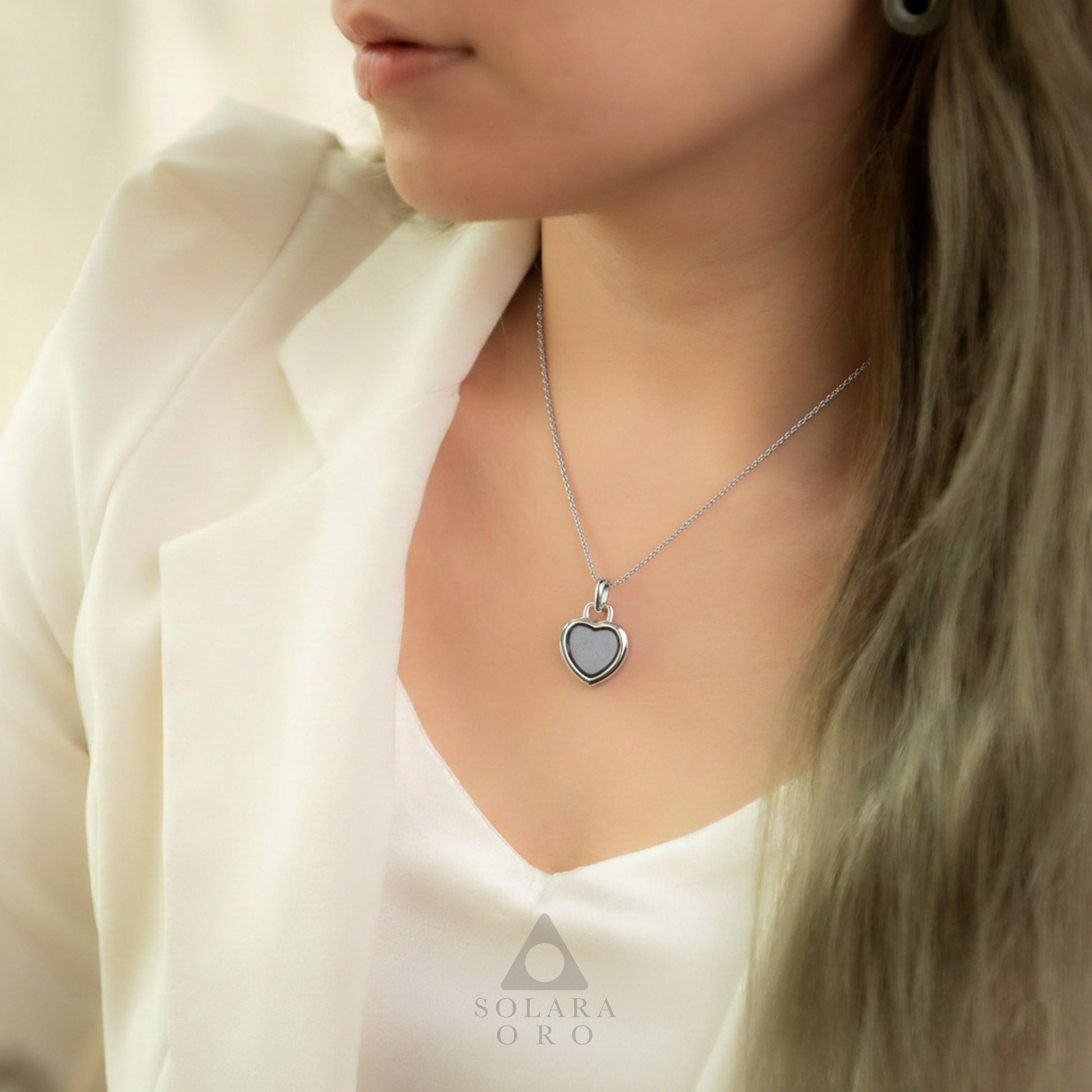 Woman wearing a silver necklace with a heart-shaped pendant, branded 'Solara Oro'.