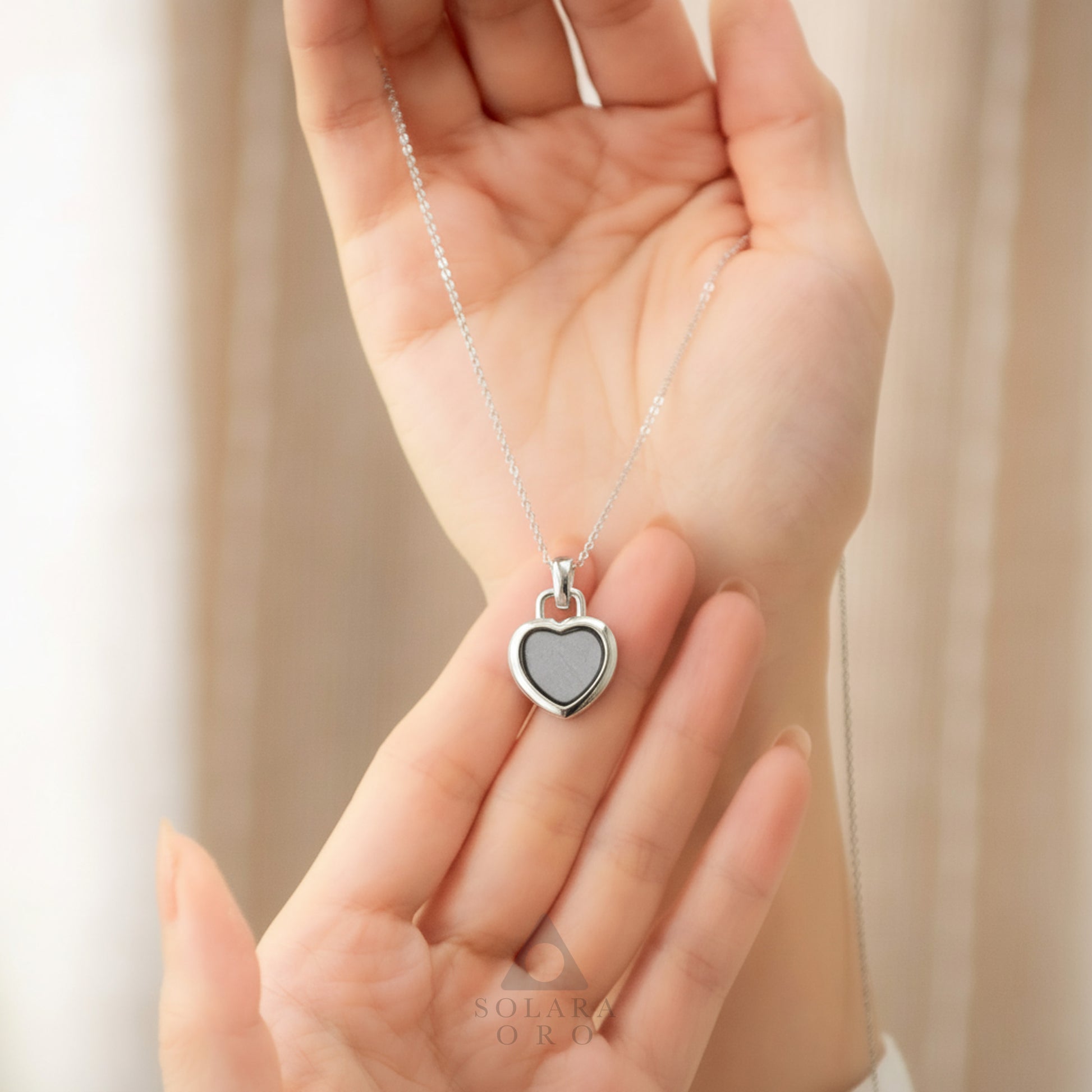 Heart-shaped silver pendant necklace on a chain held in a hand against a neutral background.