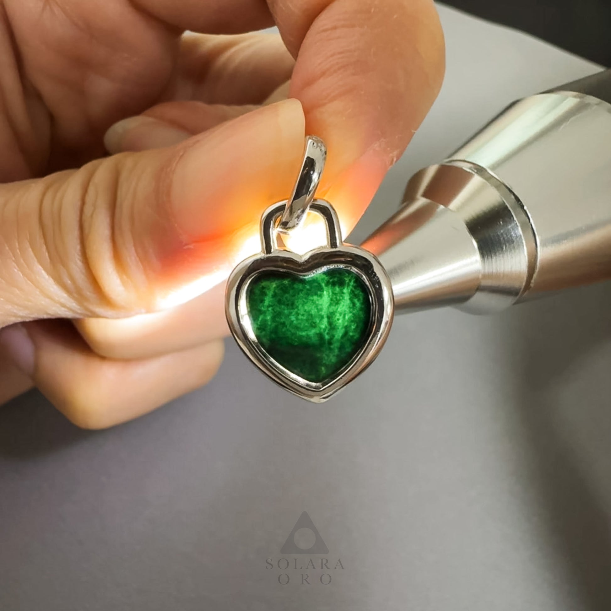 Heart-shaped pendant with green center held by a hand against a neutral background