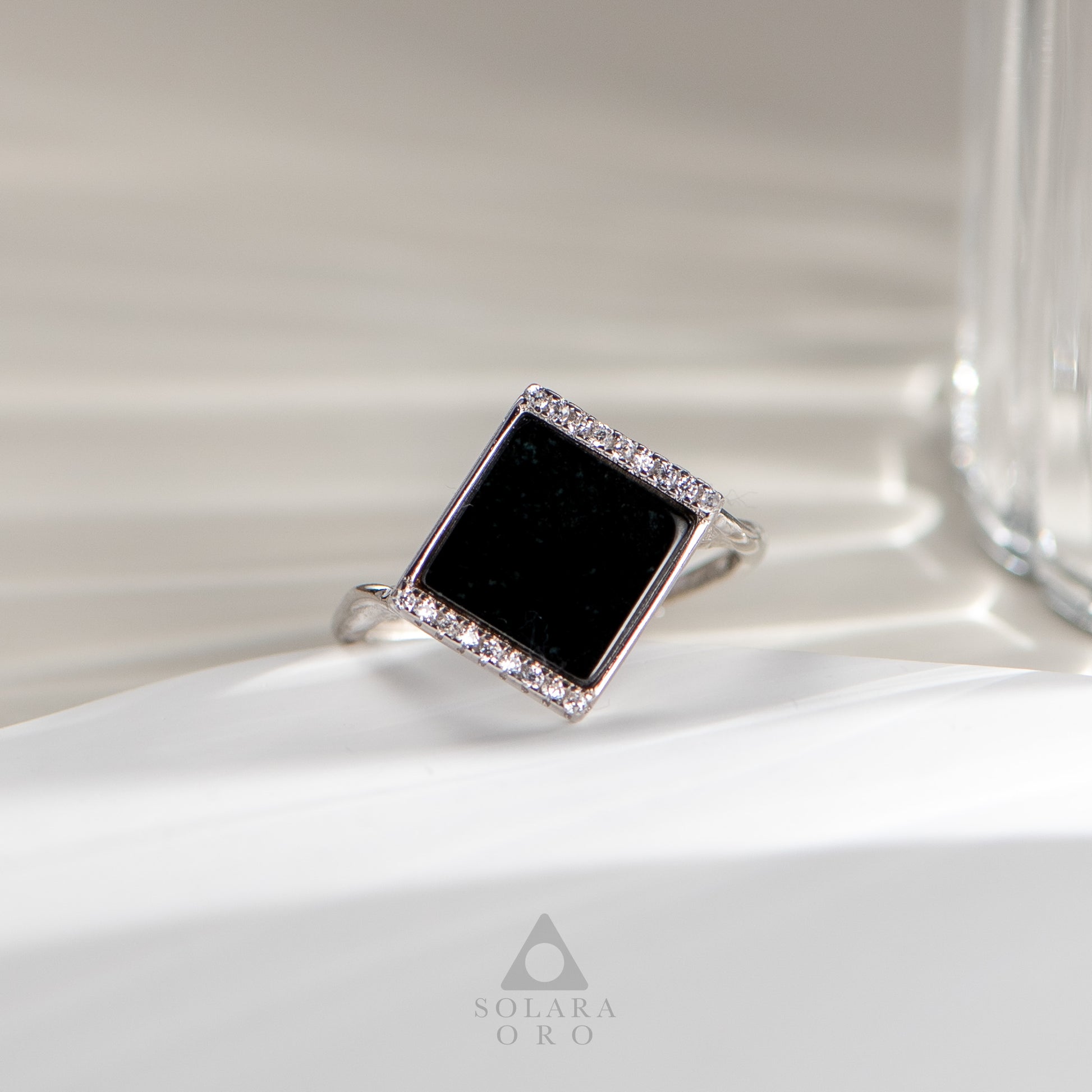 Silver ring with a black square stone and diamond accents on a white surface, brand 'Soleara Oro' visible.