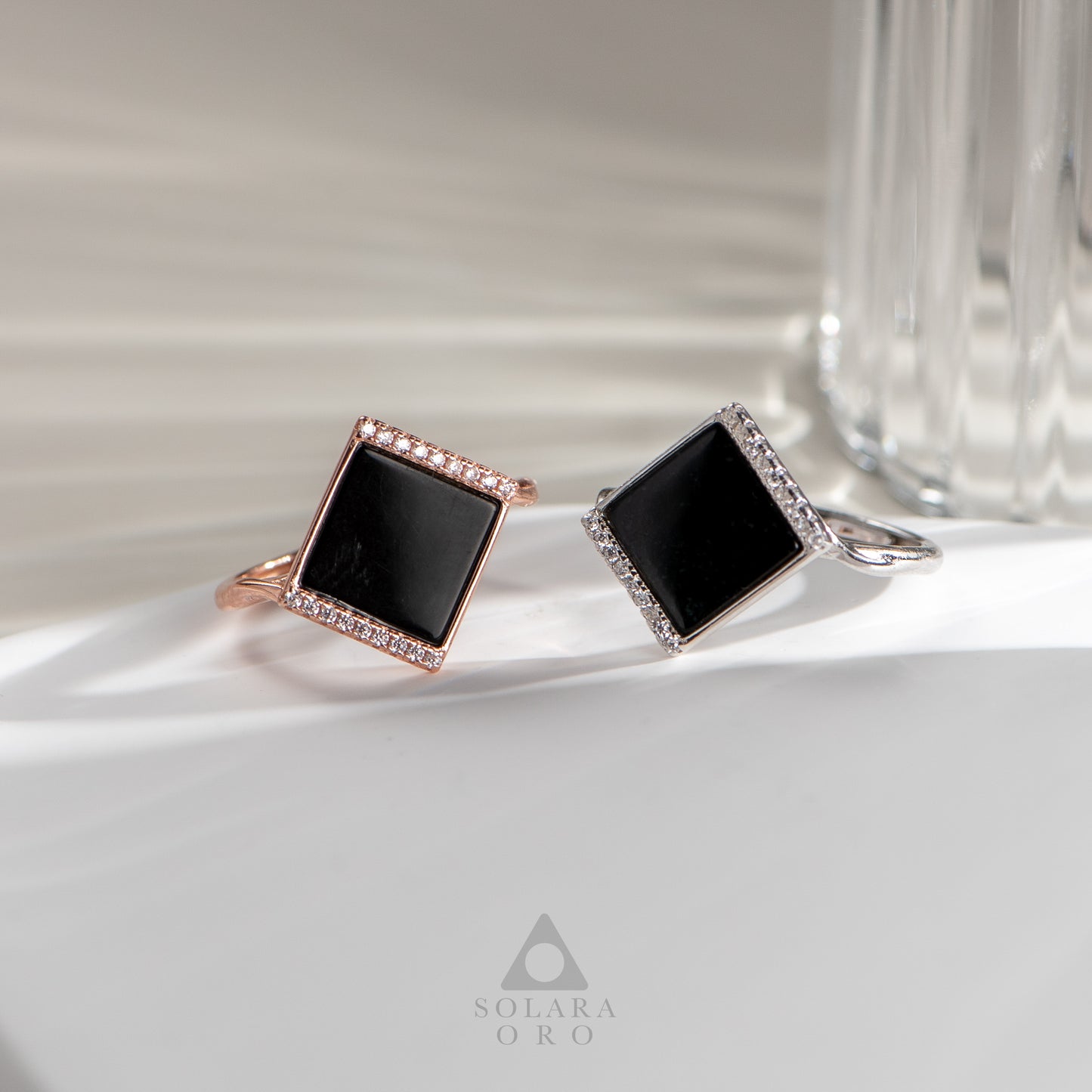 Two square black gemstone rings, one rose gold and one silver, on a light surface with a glass object in the background.