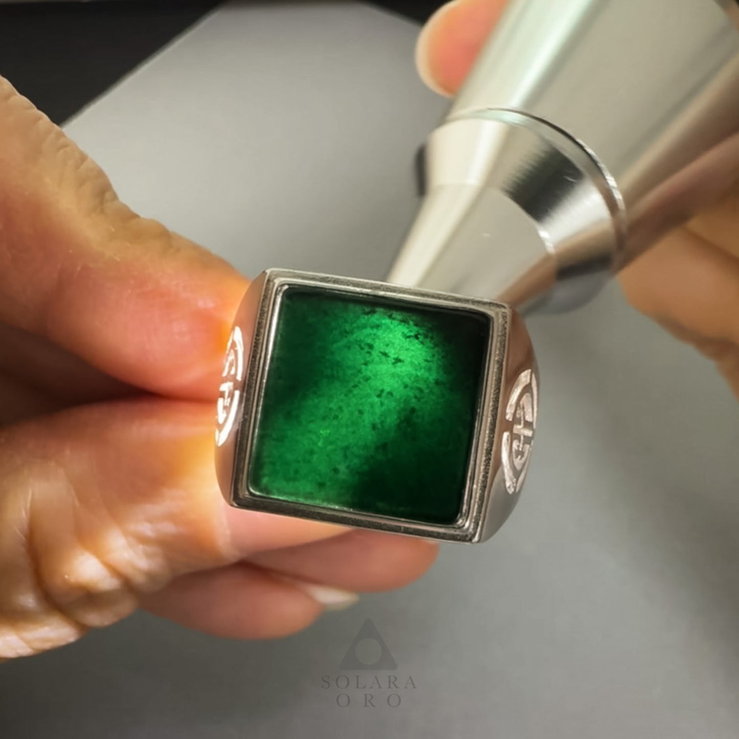 Close-up of a hand holding a metallic object with a green gemstone, branded 'Solaire Oro'.