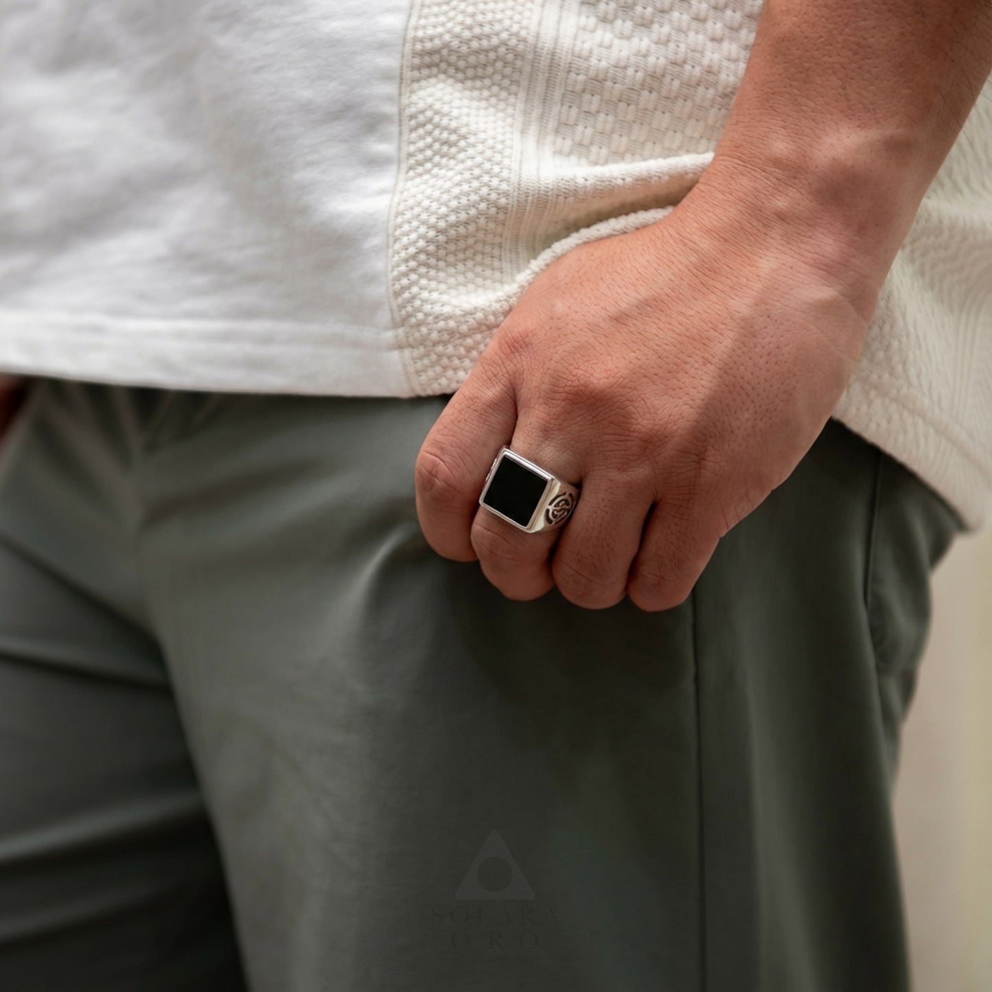 Person wearing a white textured shirt and green pants with a black ring on a neutral background