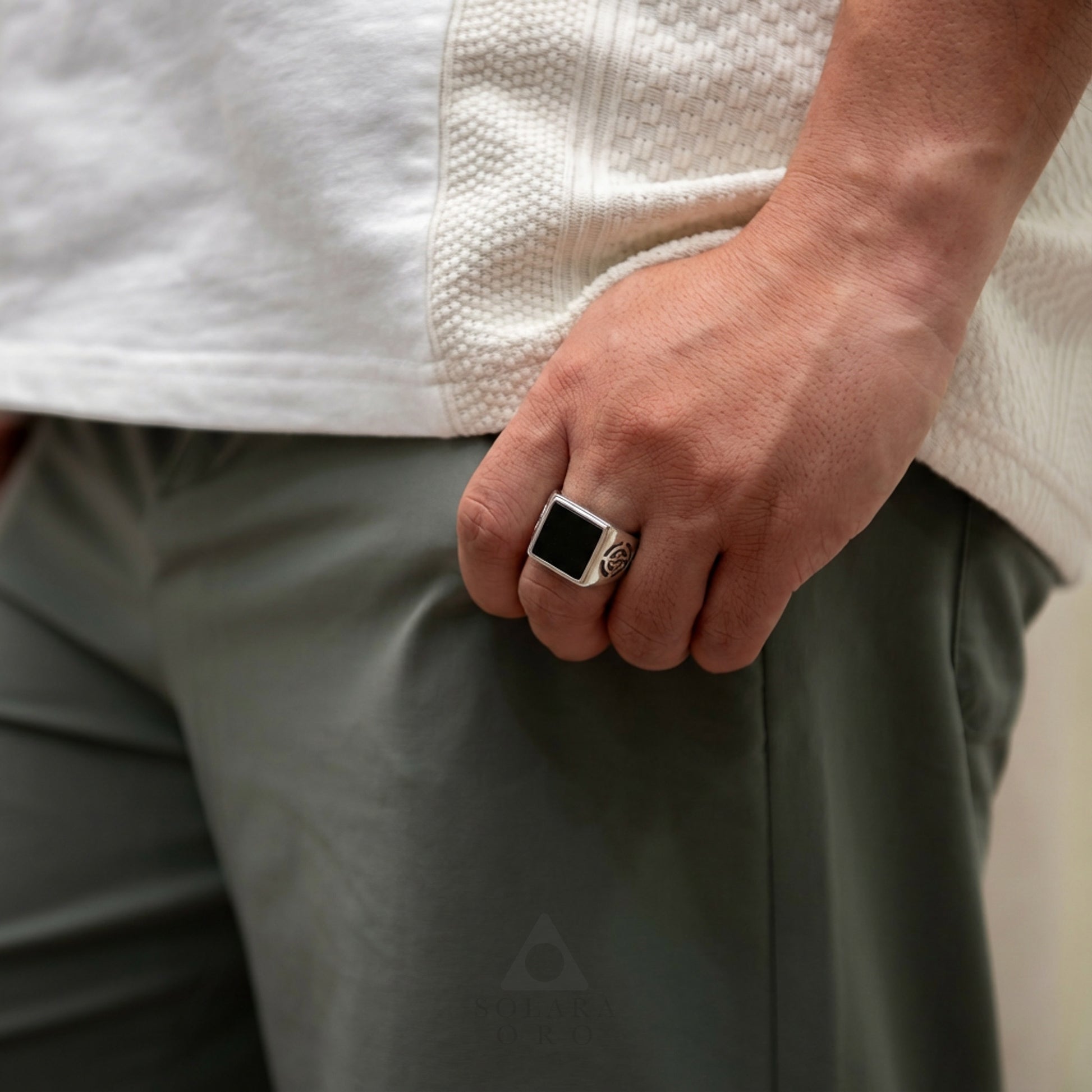 Person wearing a white textured shirt and green pants with a black ring on a neutral background