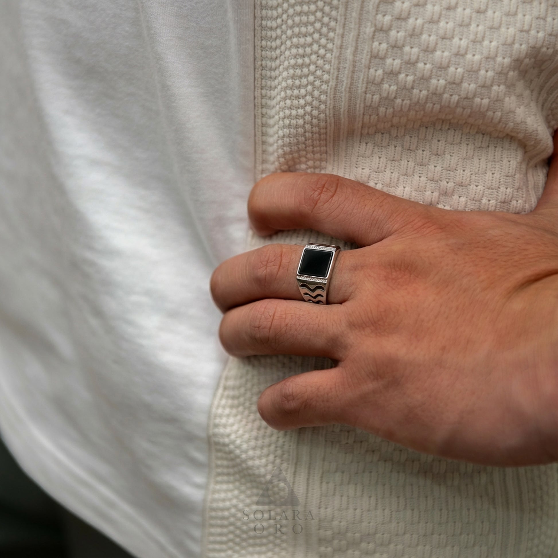 Hand wearing a silver ring with a black stone, against a textured white background