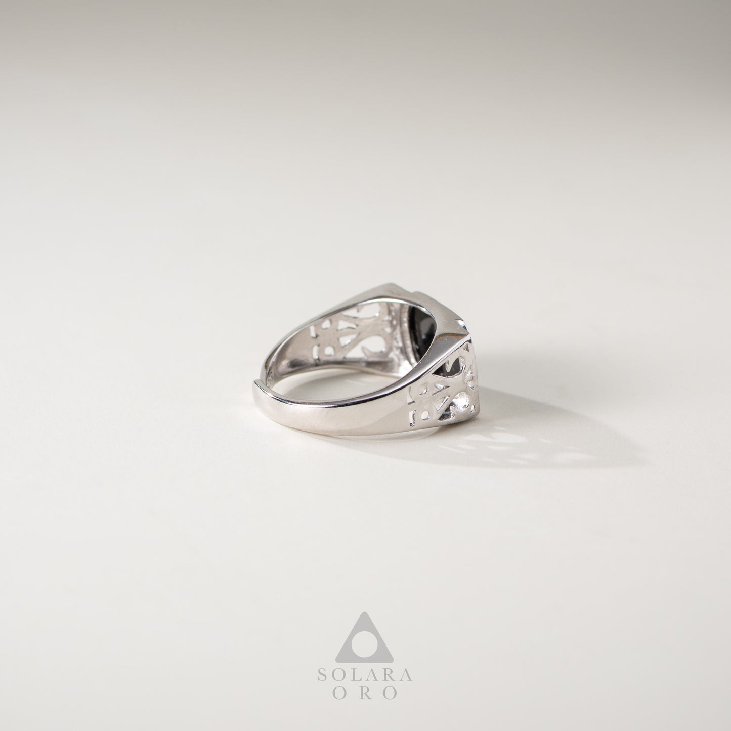 Silver ring with intricate design on a light gray background, featuring the Solara Oro logo.