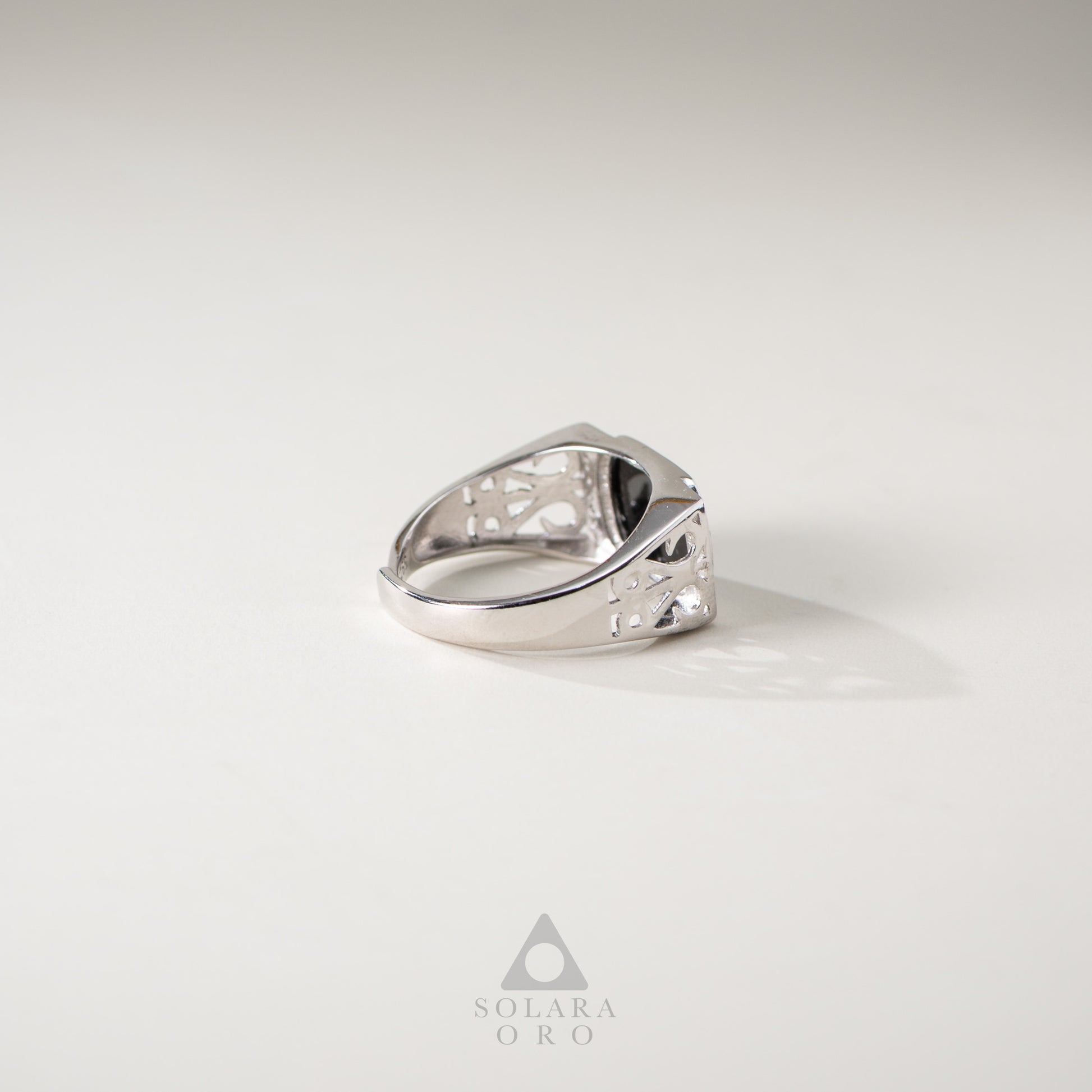 Silver ring with intricate design on a light gray background, featuring the Solara Oro logo.