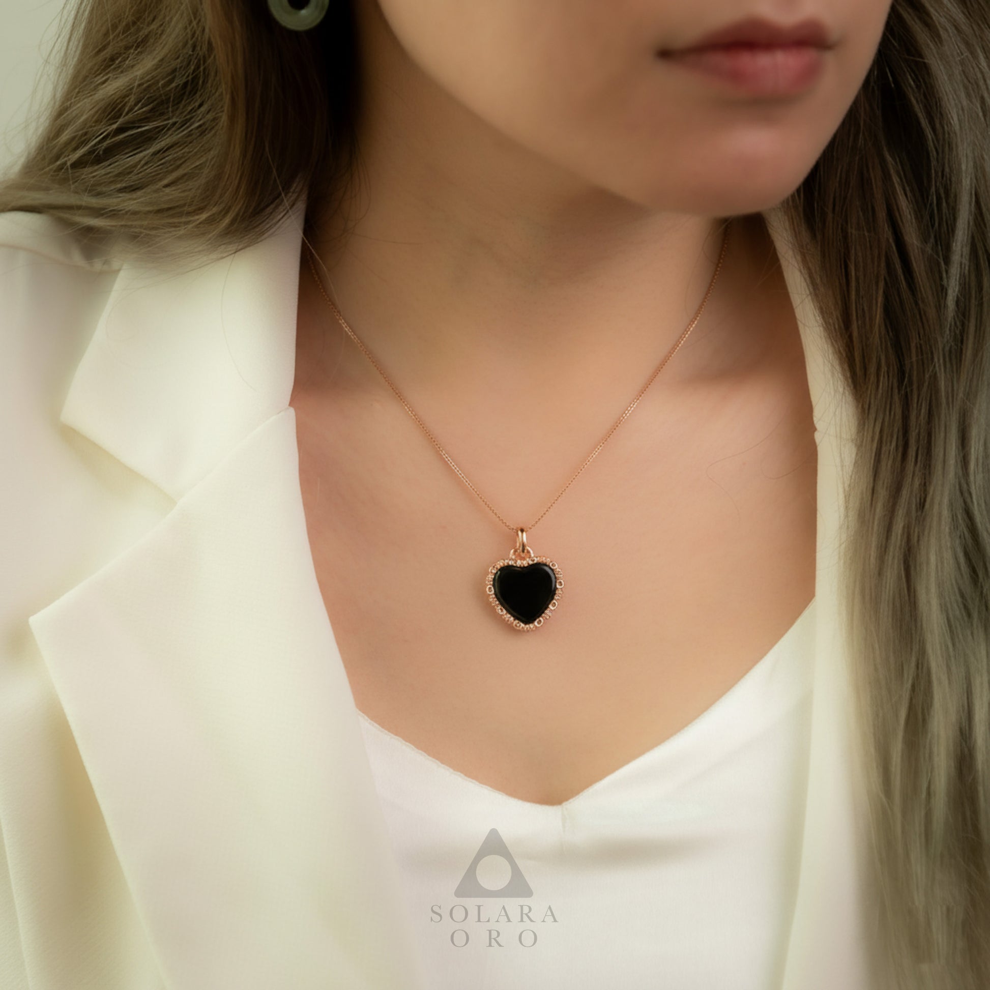 Close-up of a person wearing a necklace with a black heart pendant, branded 'Solaara Oro'.