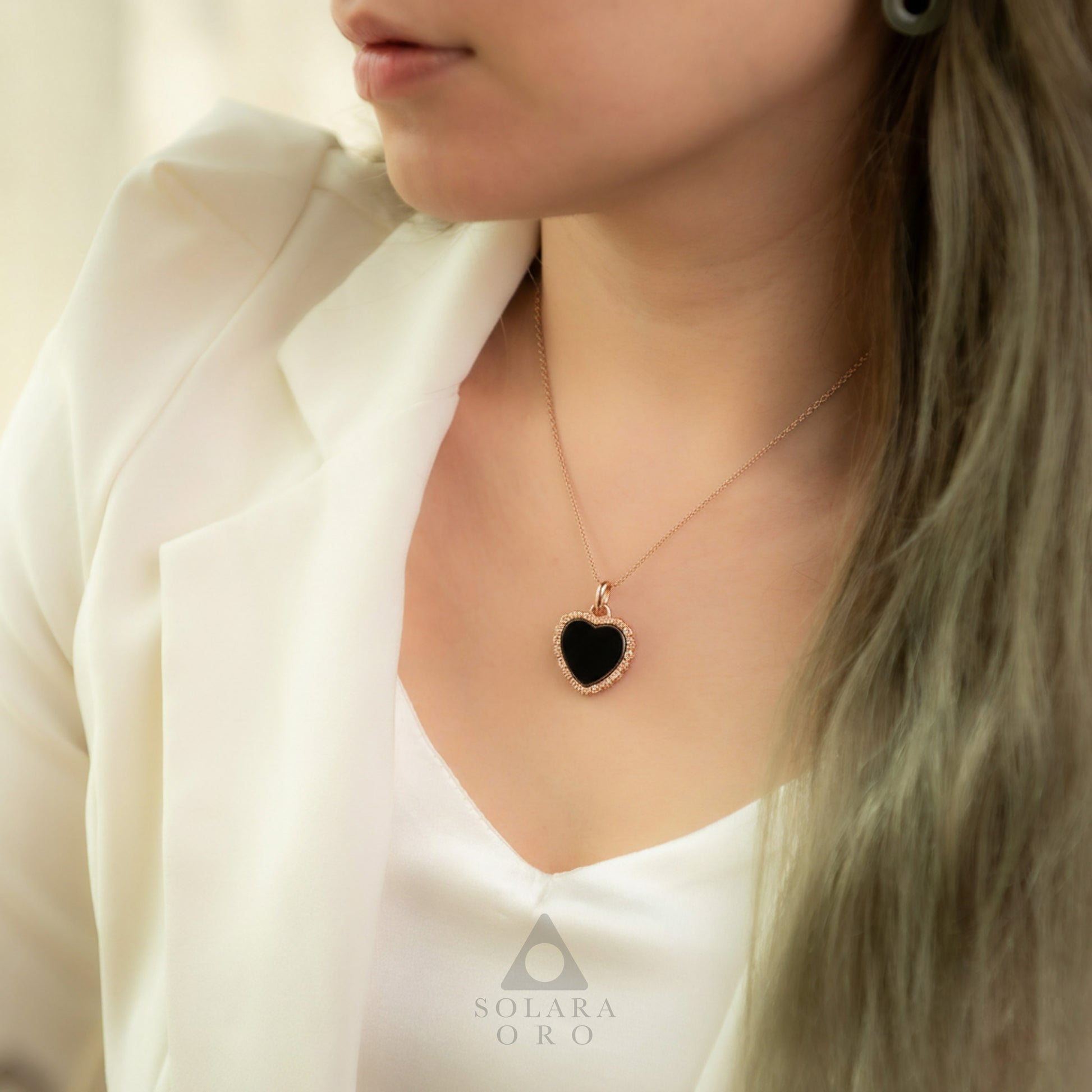 Person wearing a necklace with a black heart pendant, brand 'Solerara Oro' visible.