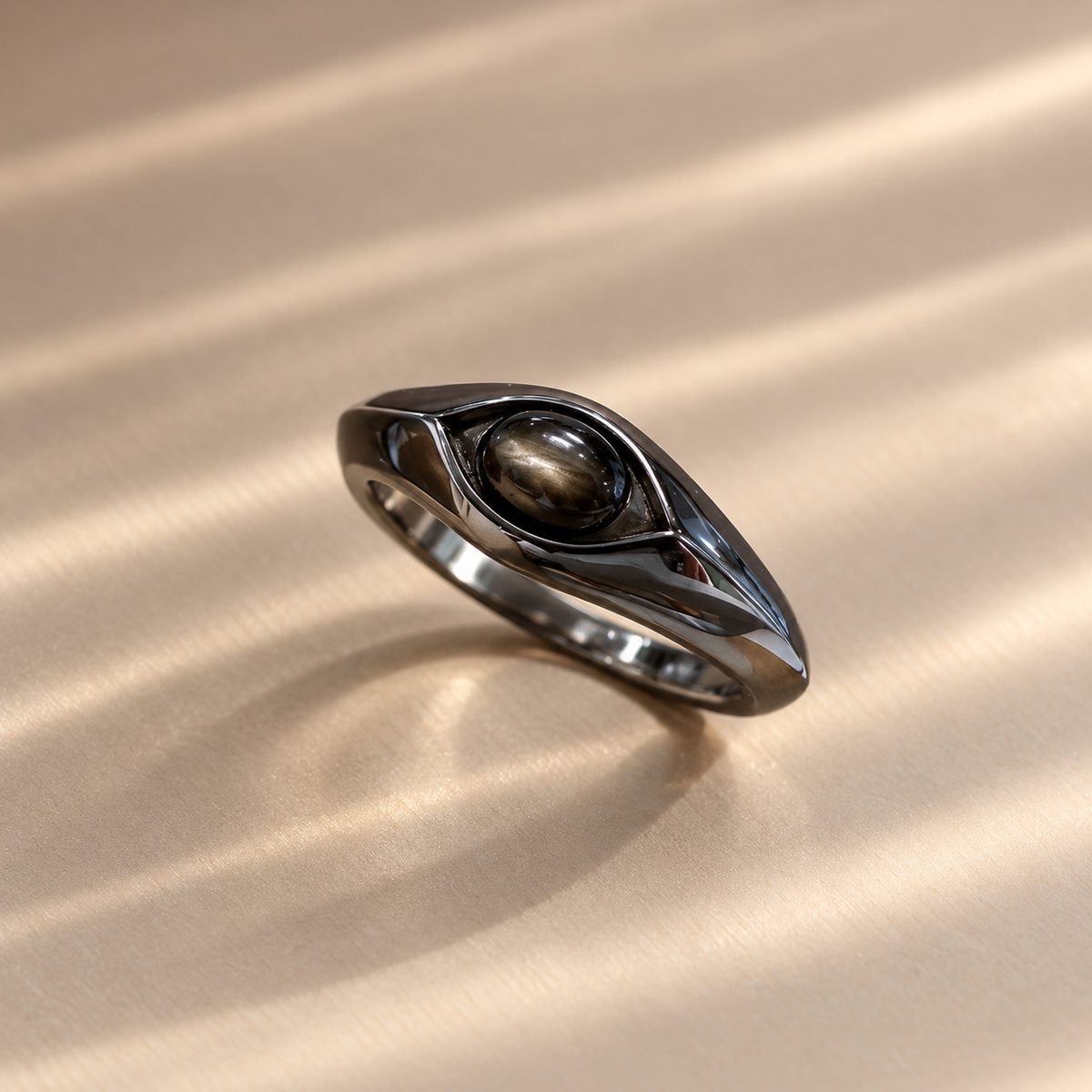 A polished dark metallic ring with a central oval brown gemstone, resting on a textured beige surface with soft diagonal shadows.