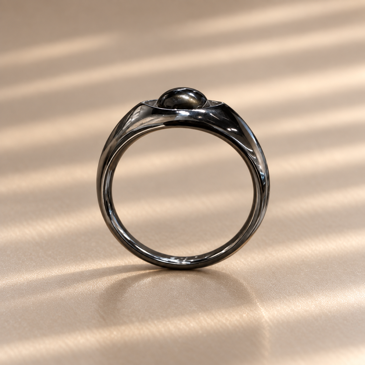 A polished dark metallic ring with a unique dome-like top detail, resting on a textured beige surface with soft diagonal shadows.