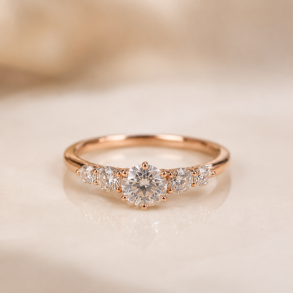 A rose gold engagement ring with a round-cut center diamond and smaller side diamonds, displayed on a soft beige surface.