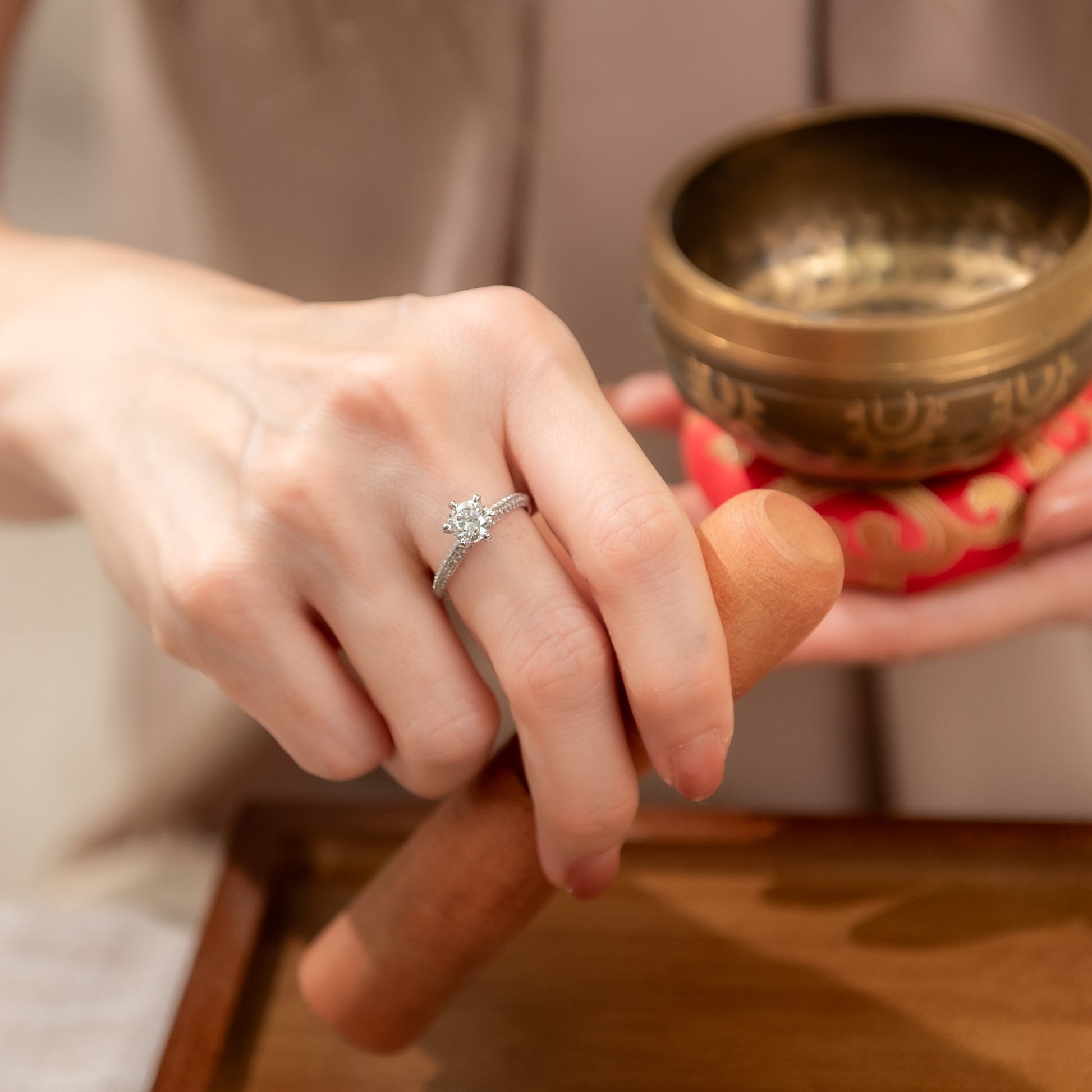 Hand wearing a diamond ring with a blurred background of a person holding a brass bowl.