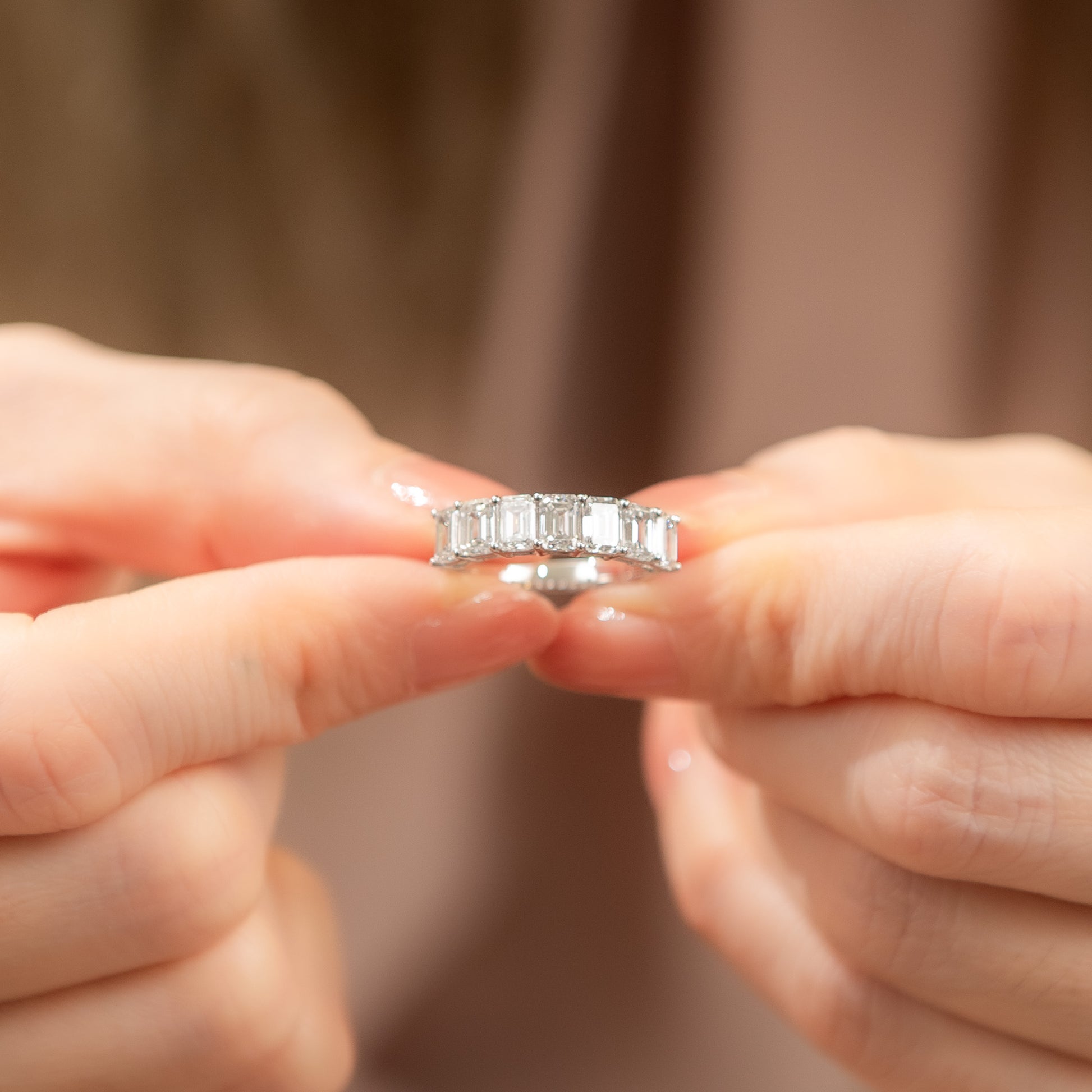 Silver ring with emerald-cut gemstones held between two hands against a neutral background