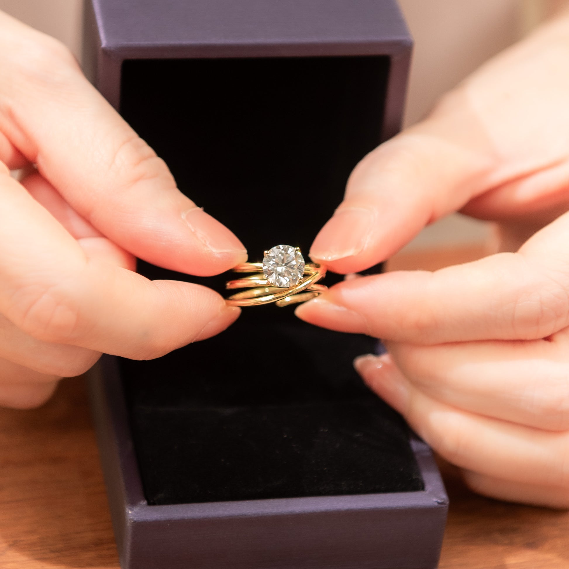 Gold ring with a diamond held by hands against a dark purple box.